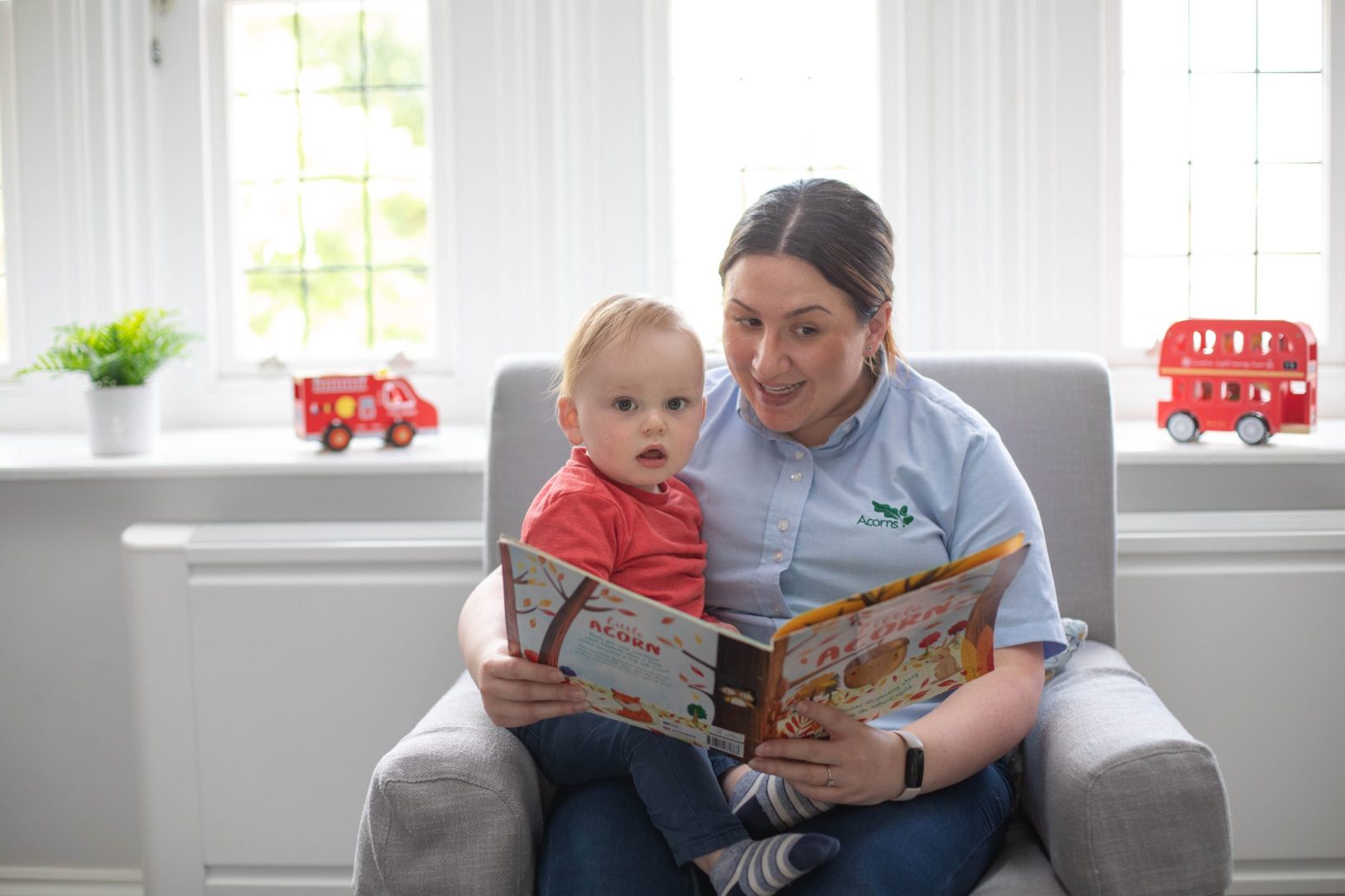 Acorns Nurseries An adult and a child sit on a chair, sharing the joy of a picture book in a well-lit room. Toy buses rest on the window sill, adding to this caring moment of childcare and connection.