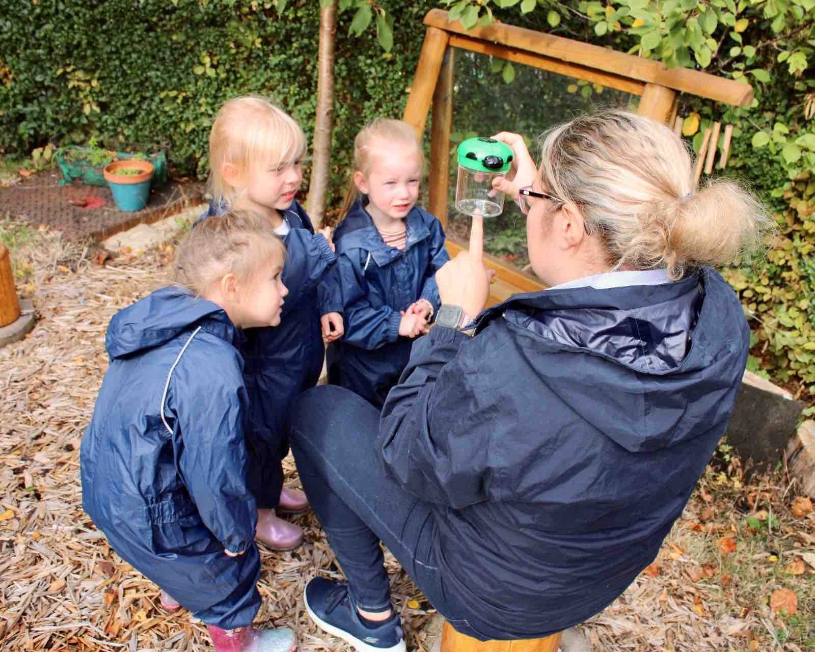 Acorns Nurseries A Forest School teacher shows a green object to three young children in blue raincoats, sparking curiosity as they explore the wonders of nature together.