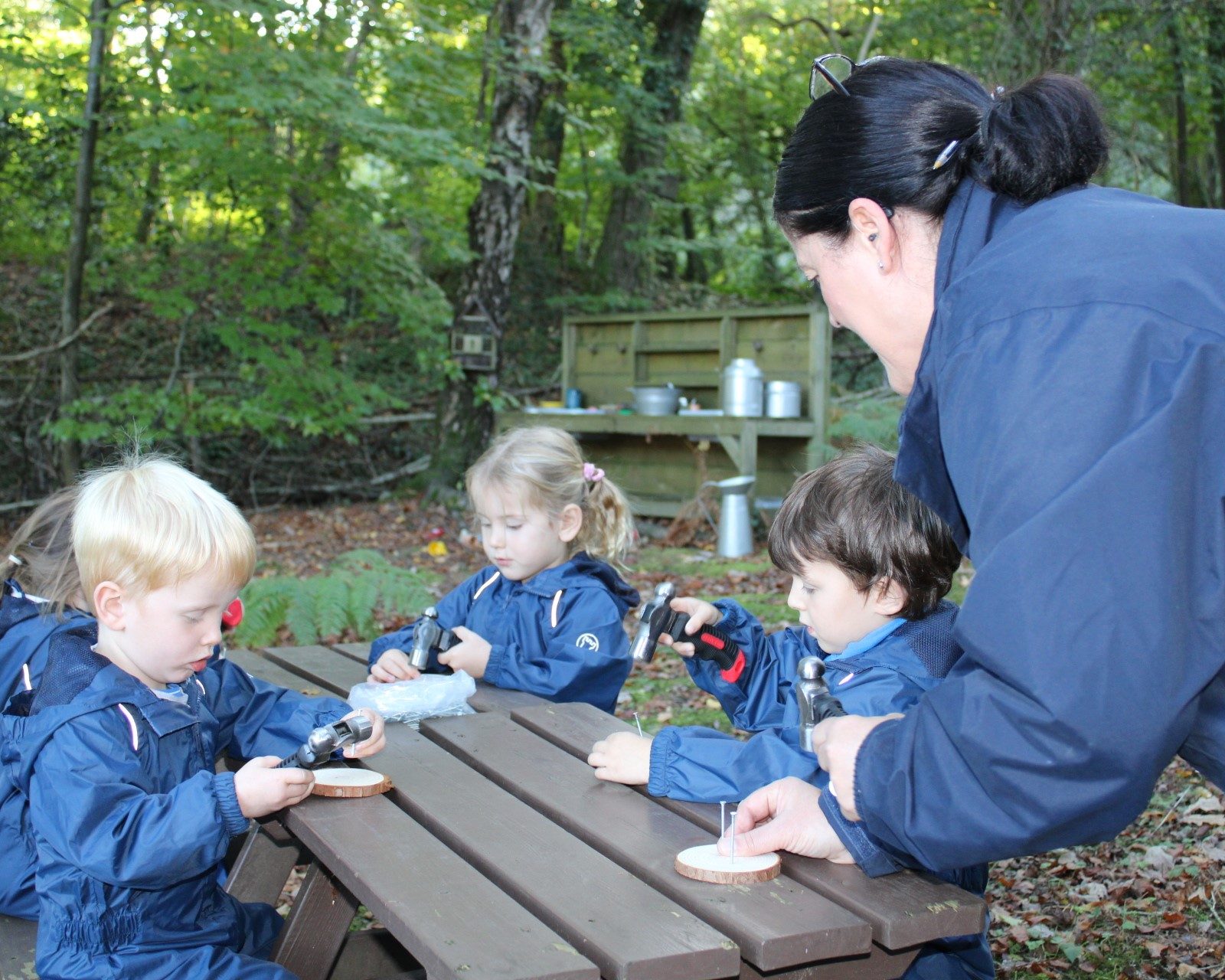 Acorns Nurseries Children and an adult sit at a picnic table in a forested area, immersed in a Forest School craft activity with small wooden pieces, showcasing the essence of outdoor education and nature learning.