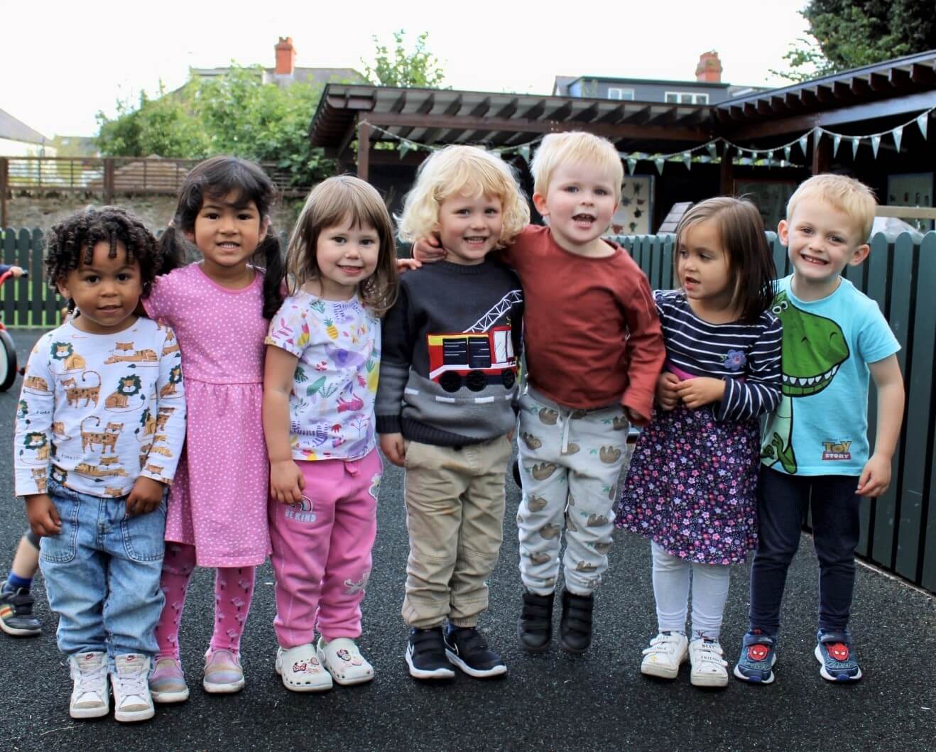 Acorns Nurseries Seven children stand in a row, beaming with joy outdoors, as if in their second home, surrounded by playground equipment and trees in the background.