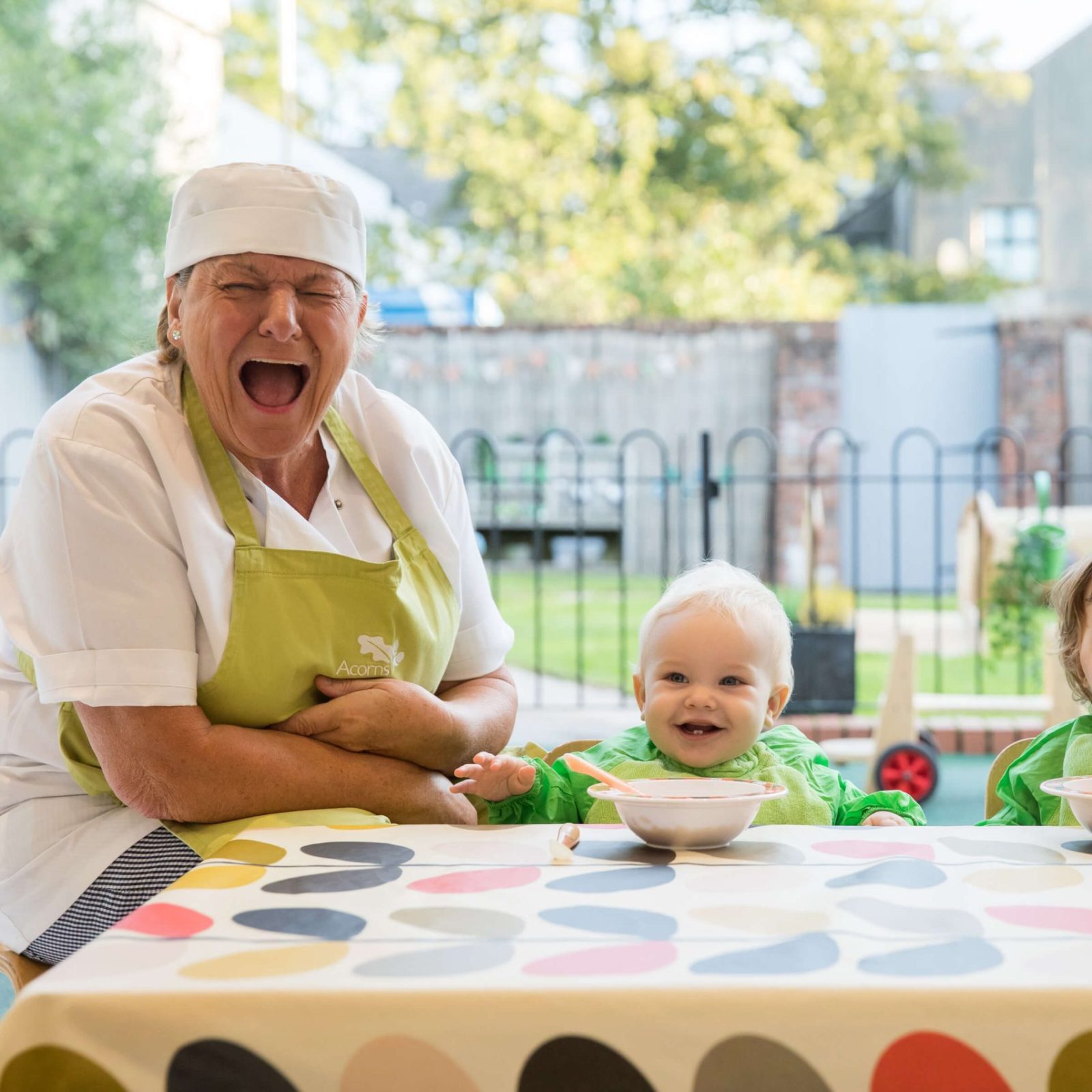 Acorns Nurseries A smiling woman in a chef's hat sits beside two toddlers at a cozy outdoor table, complete with bowls of food and a colorful tablecloth, creating a warm home atmosphere.