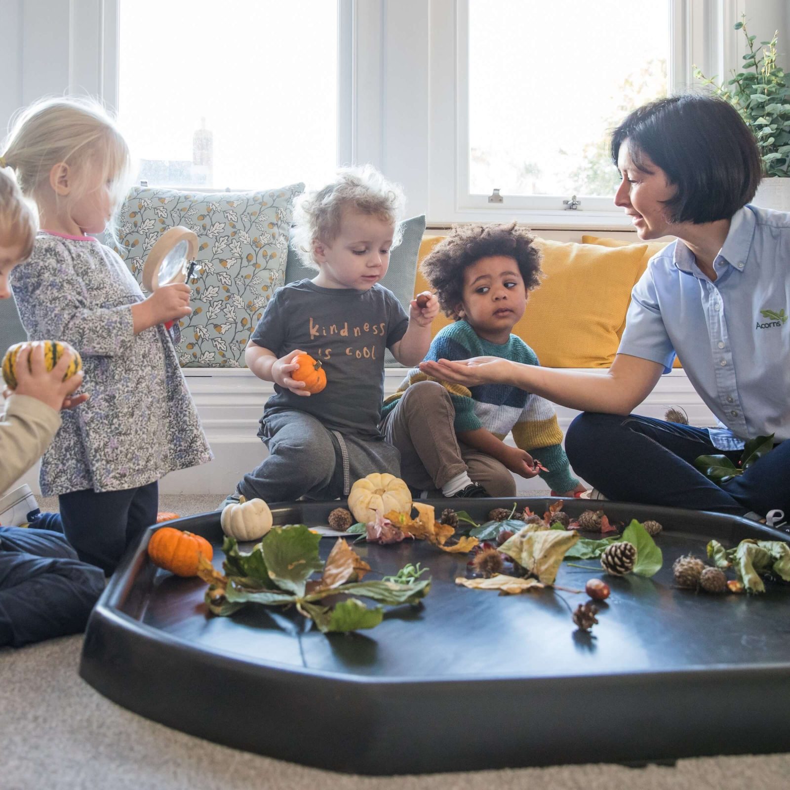 Acorns Nurseries Children and a teacher at home explore autumn leaves and small pumpkins on a tray indoors, fostering a cozy atmosphere of learning and discovery.