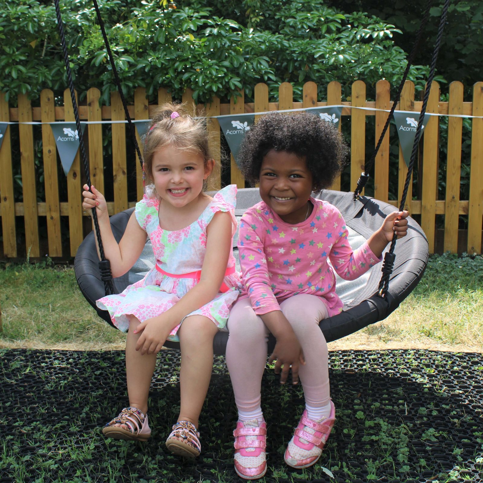 Acorns Nurseries Two children sit together on a swing in a garden at home, smiling at the camera, with trees and a wooden fence in the background.