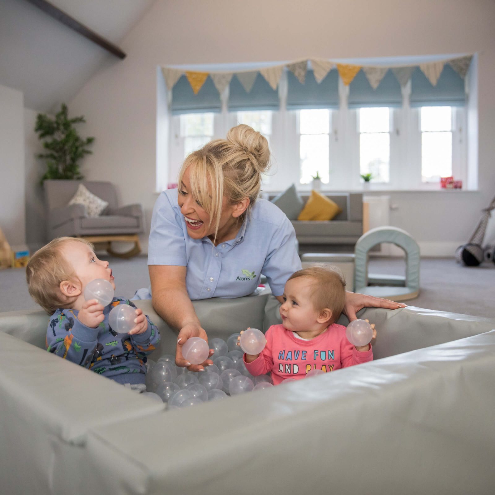 Acorns Nurseries In a spacious, well-lit room that feels just like home, a caregiver and two toddlers joyfully play with balls in the soft play area, with a charming teepee in the background.