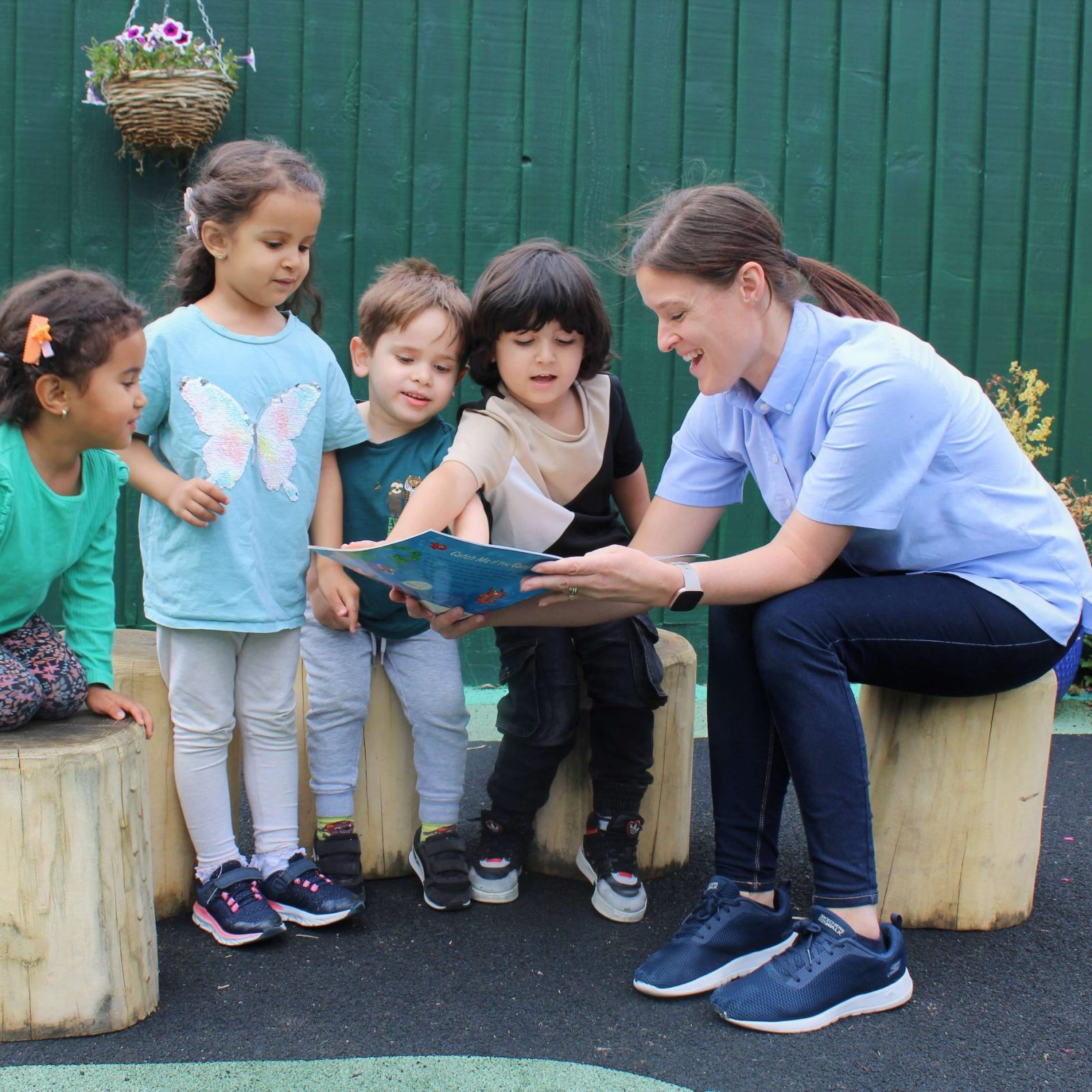 Acorns Nurseries A teacher reads a book to four young children seated on tree stump seats, creating a cozy outdoor classroom with a green fence backdrop that feels like home.