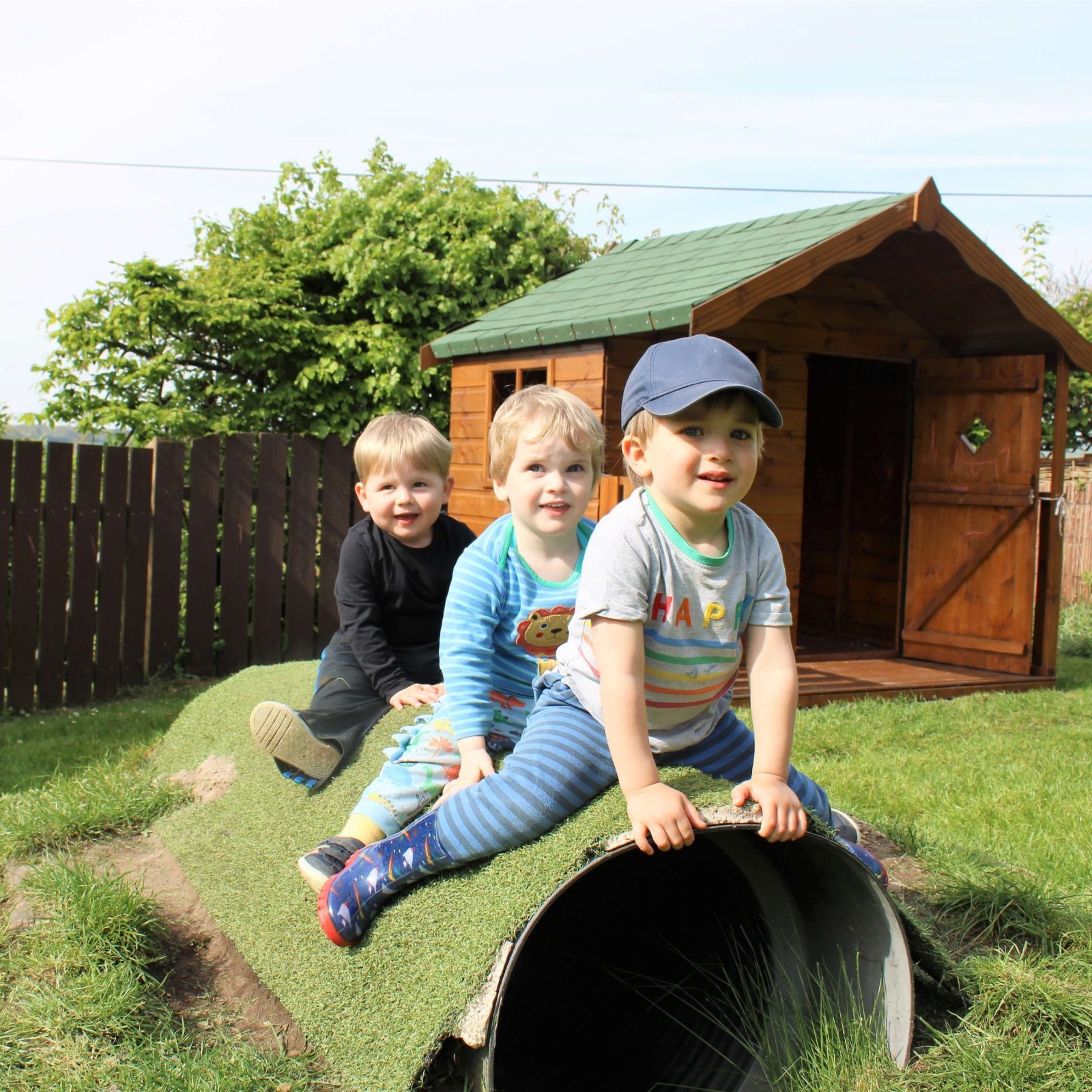 Acorns Nurseries Three children sitting and smiling on a grass-covered tunnel in a garden, with their cozy wooden playhouse in the background, evoking a sense of home.