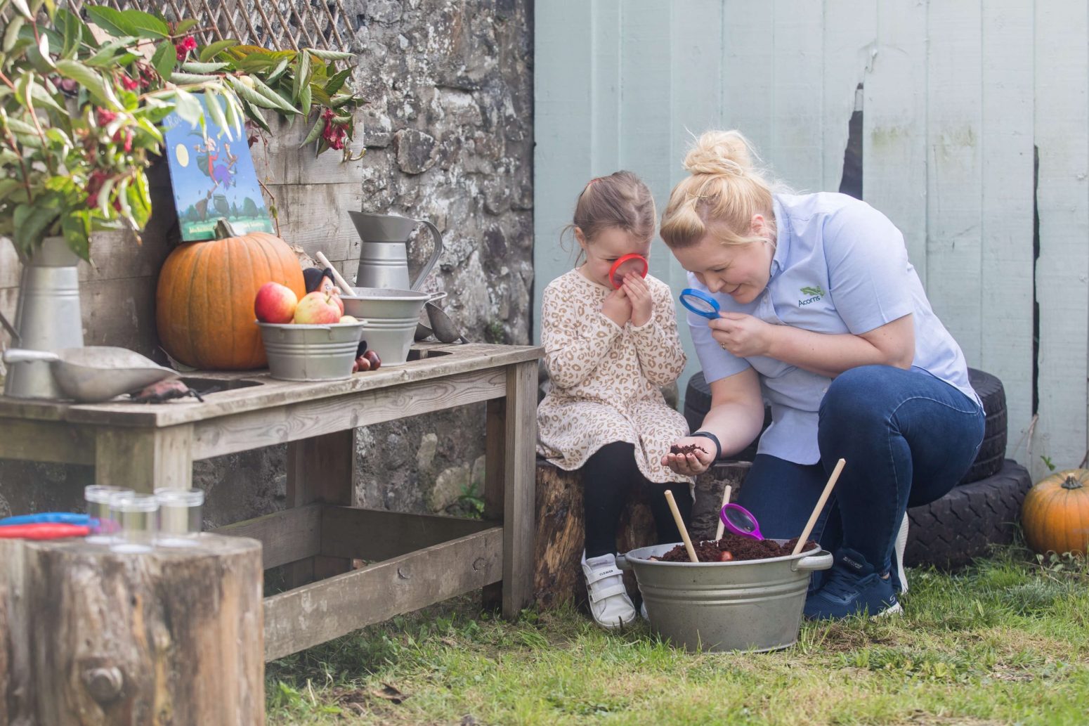 Acorns Nurseries In a cozy garden setting, an adult and child use magnifying glasses to explore the rich soil, surrounded by pumpkins and essential gardening tools that make this home sanctuary come to life.