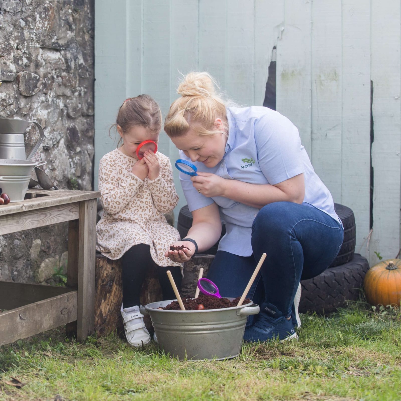 Acorns Nurseries In a cozy garden setting, an adult and child use magnifying glasses to explore the rich soil, surrounded by pumpkins and essential gardening tools that make this home sanctuary come to life.