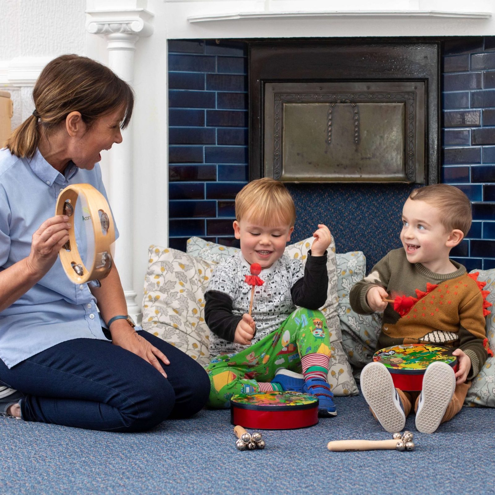 Acorns Nurseries An adult and two children sit on the floor by a cozy home fireplace, playing percussion instruments with a tambourine and toy drums.