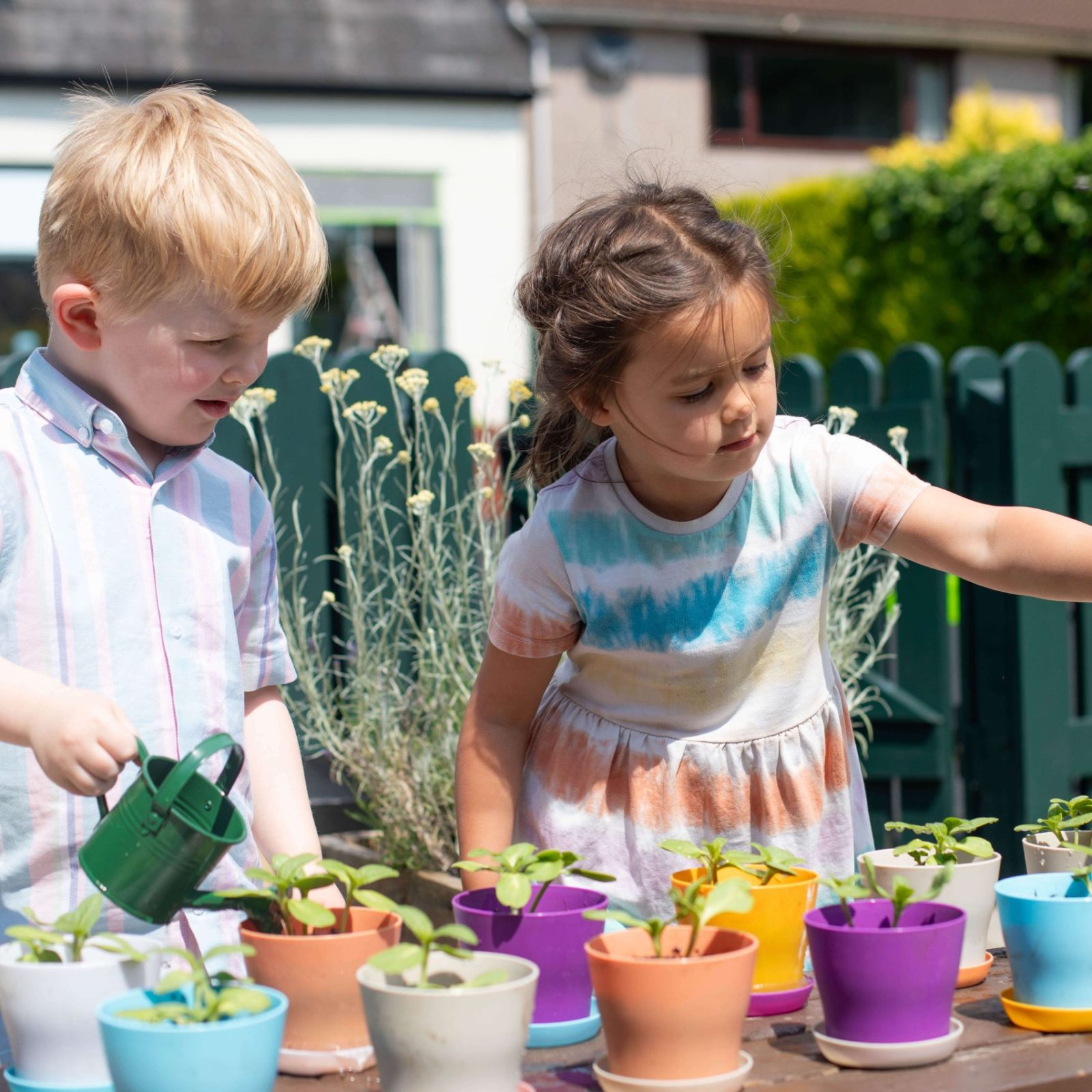 Acorns Nurseries Two children are outside in their home garden, joyfully using small watering cans to hydrate the potted plants.