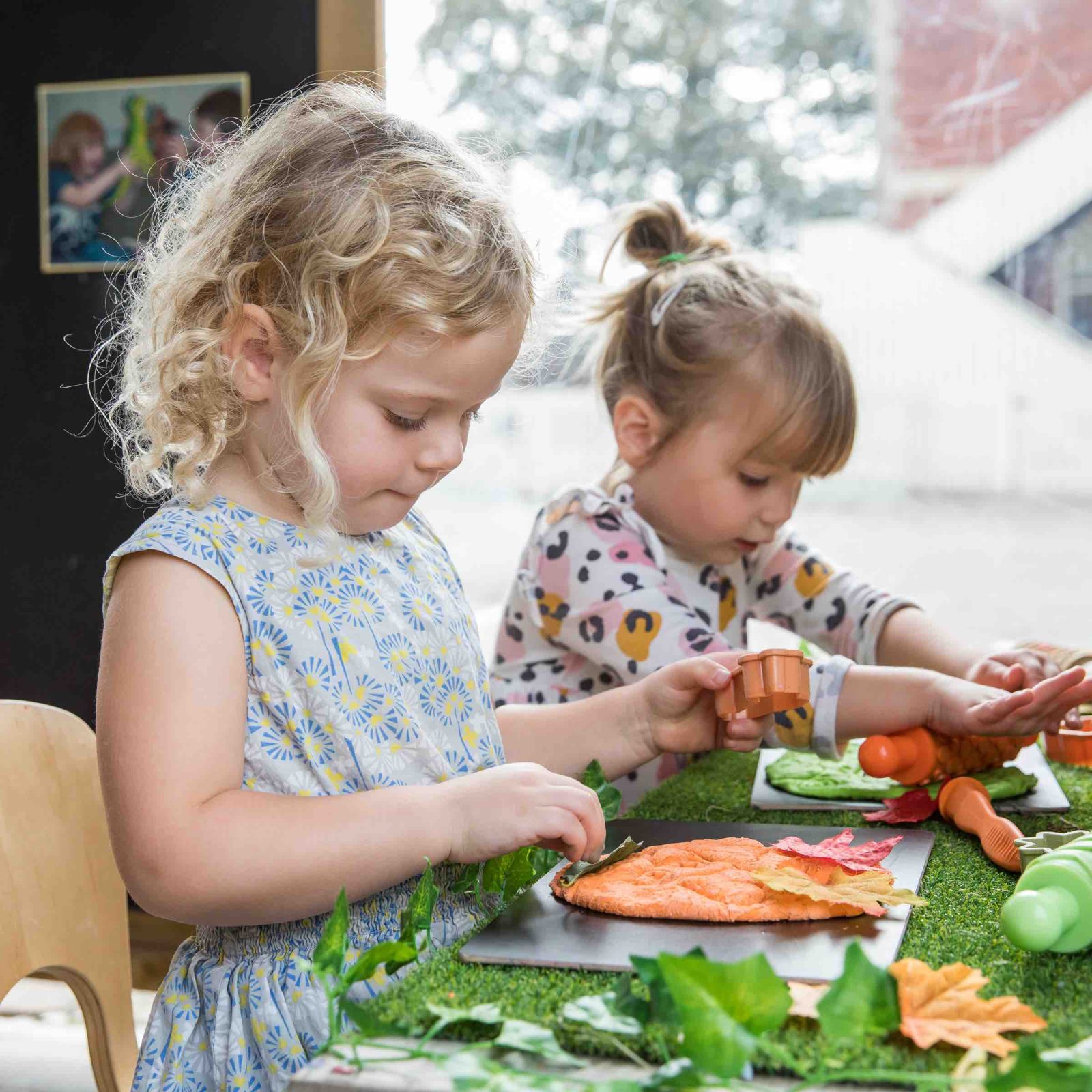 Acorns Nurseries Two young children craft with leaves and playdough at a table, bringing the essence of the Great Outdoors into their well-lit room as they focus intently on their creative activity.