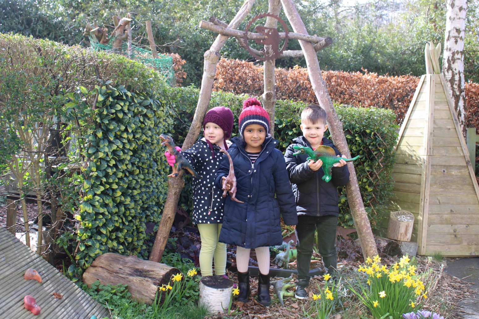 Acorns Nurseries Three children in winter clothing stand outdoors, embracing the great outdoors as they hold bird figurines, surrounded by greenery and yellow flowers.