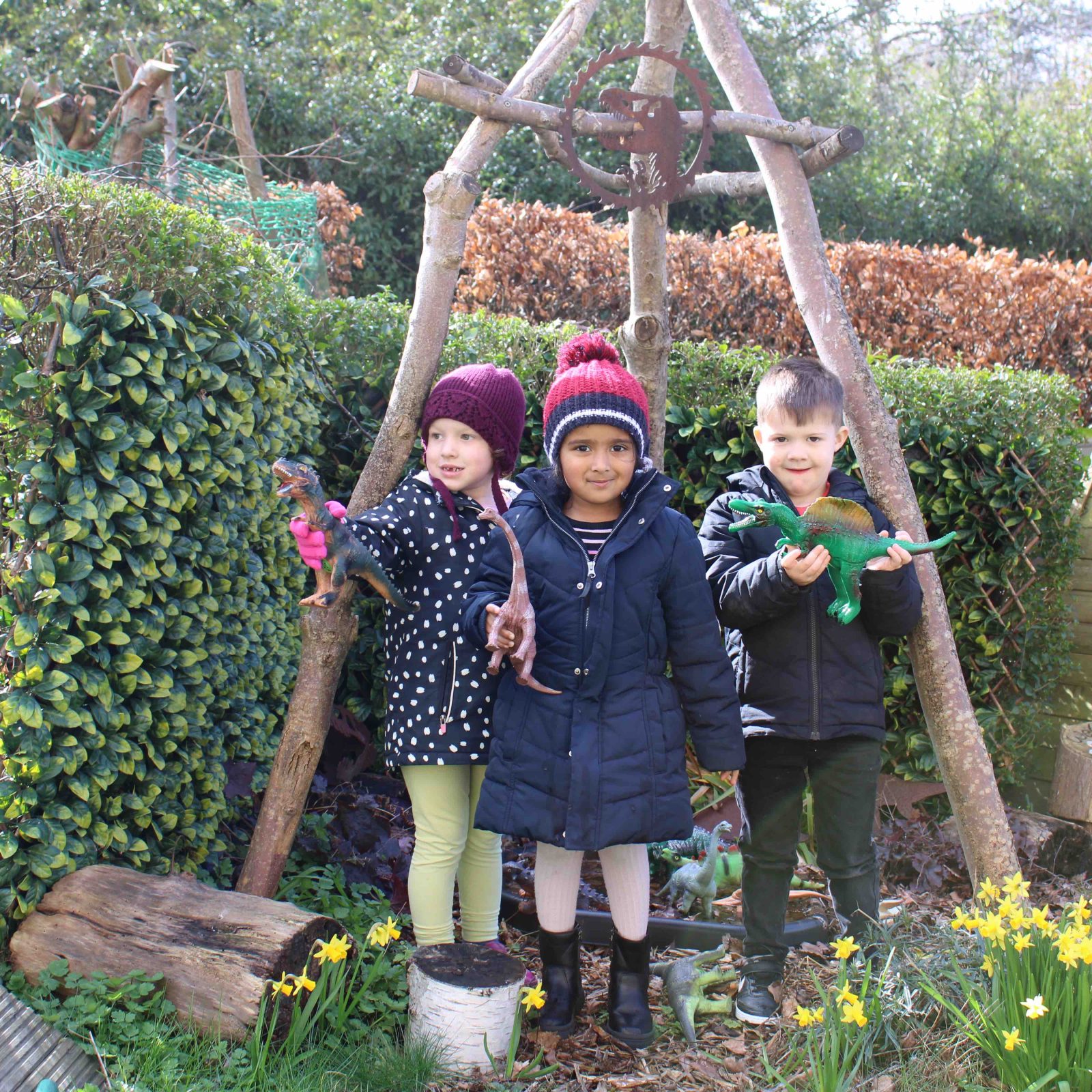 Acorns Nurseries Three children in winter clothing stand outdoors, embracing the great outdoors as they hold bird figurines, surrounded by greenery and yellow flowers.