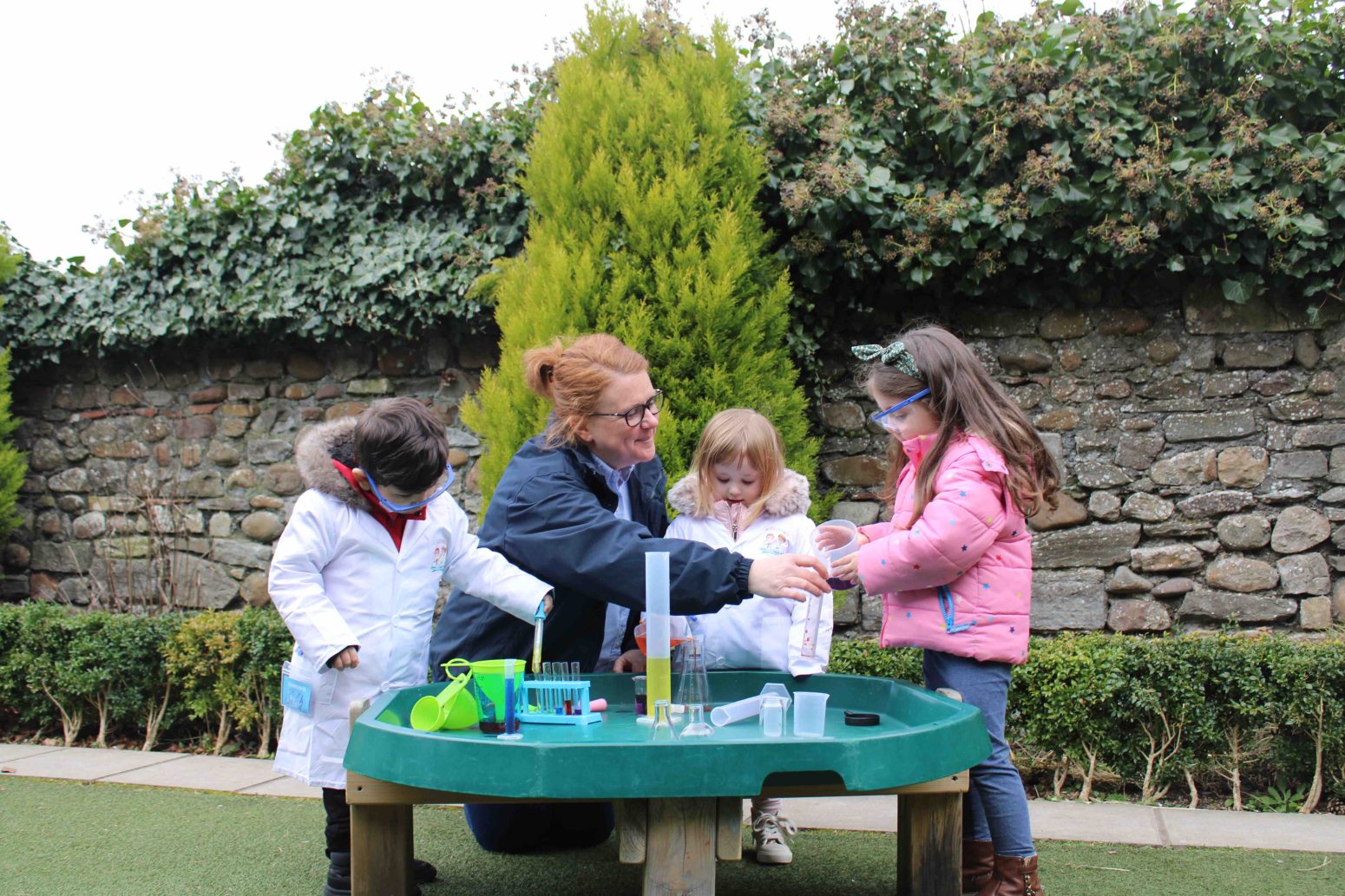 Acorns Nurseries An adult is educating three children in a science activity outdoors, where they don lab coats and use equipment on a green table.