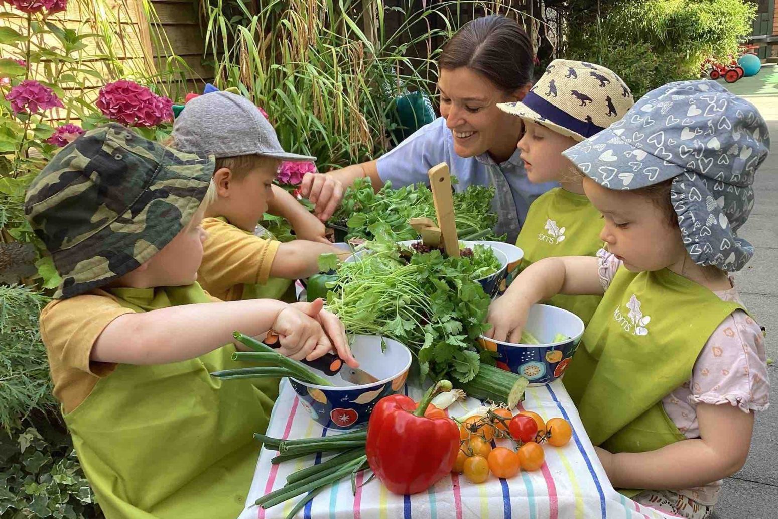 Acorns Nurseries Children wearing hats and aprons sit at a table with a caregiver, engaging in a healthy outdoor vegetable activity.