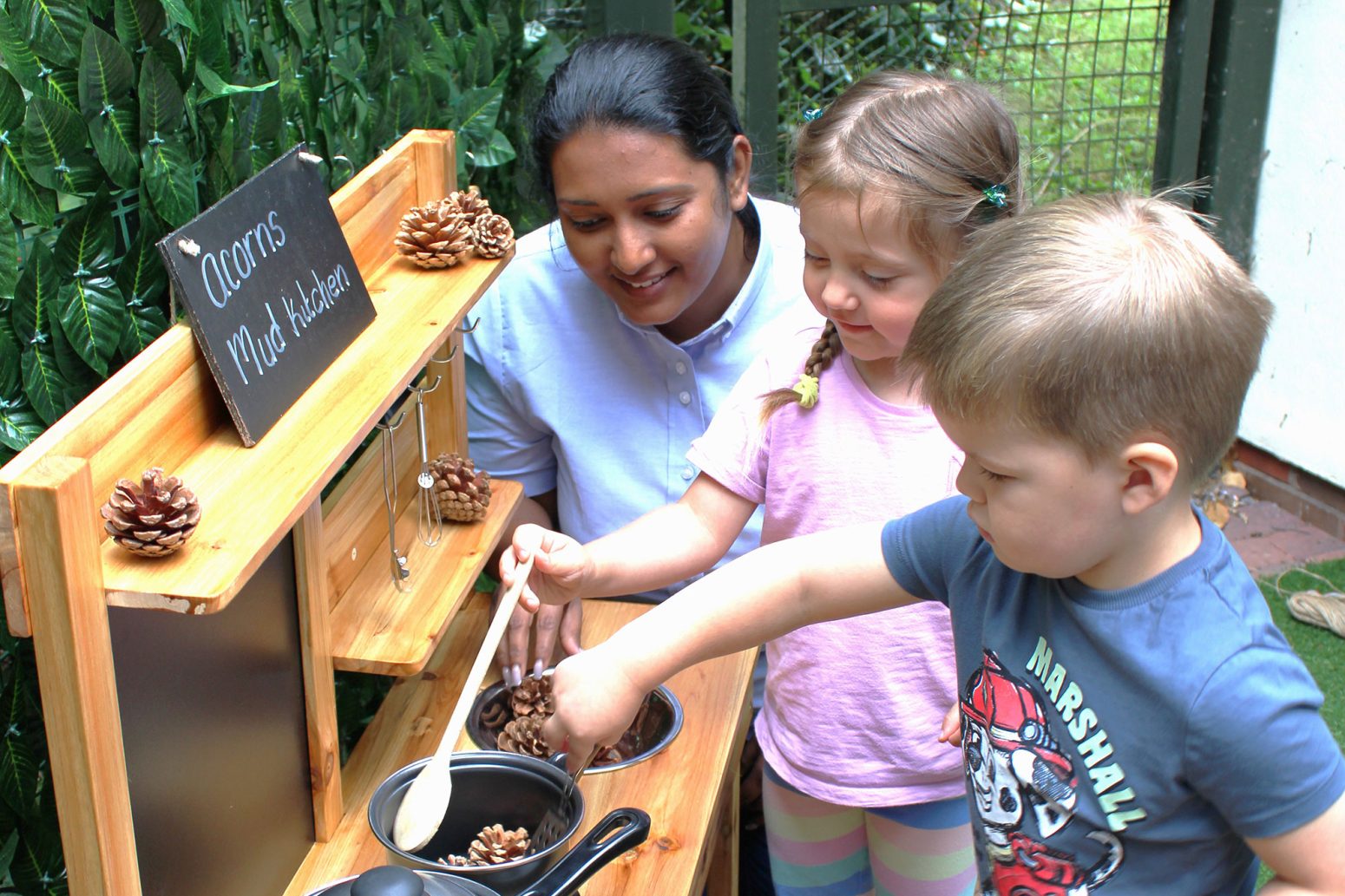 Acorns Nurseries Three children engage in a play kitchen, exploring with pine cones, while a woman carefully observes and assists them, creating an educational experience.