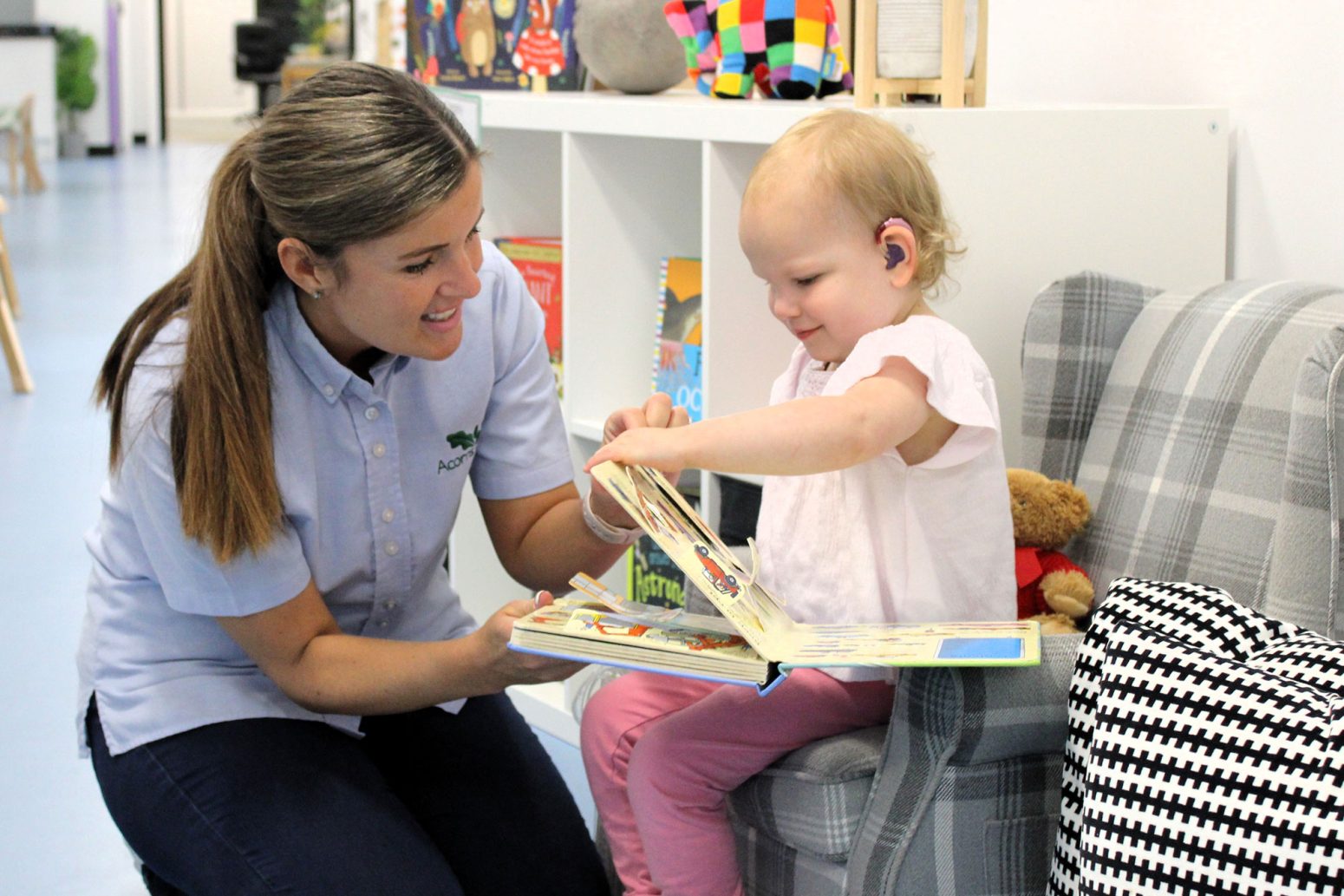 Acorns Nurseries A woman kneels beside a young child sitting on a chair, both engaged in reading a book together in the brightly lit room, fostering an atmosphere of education and care.