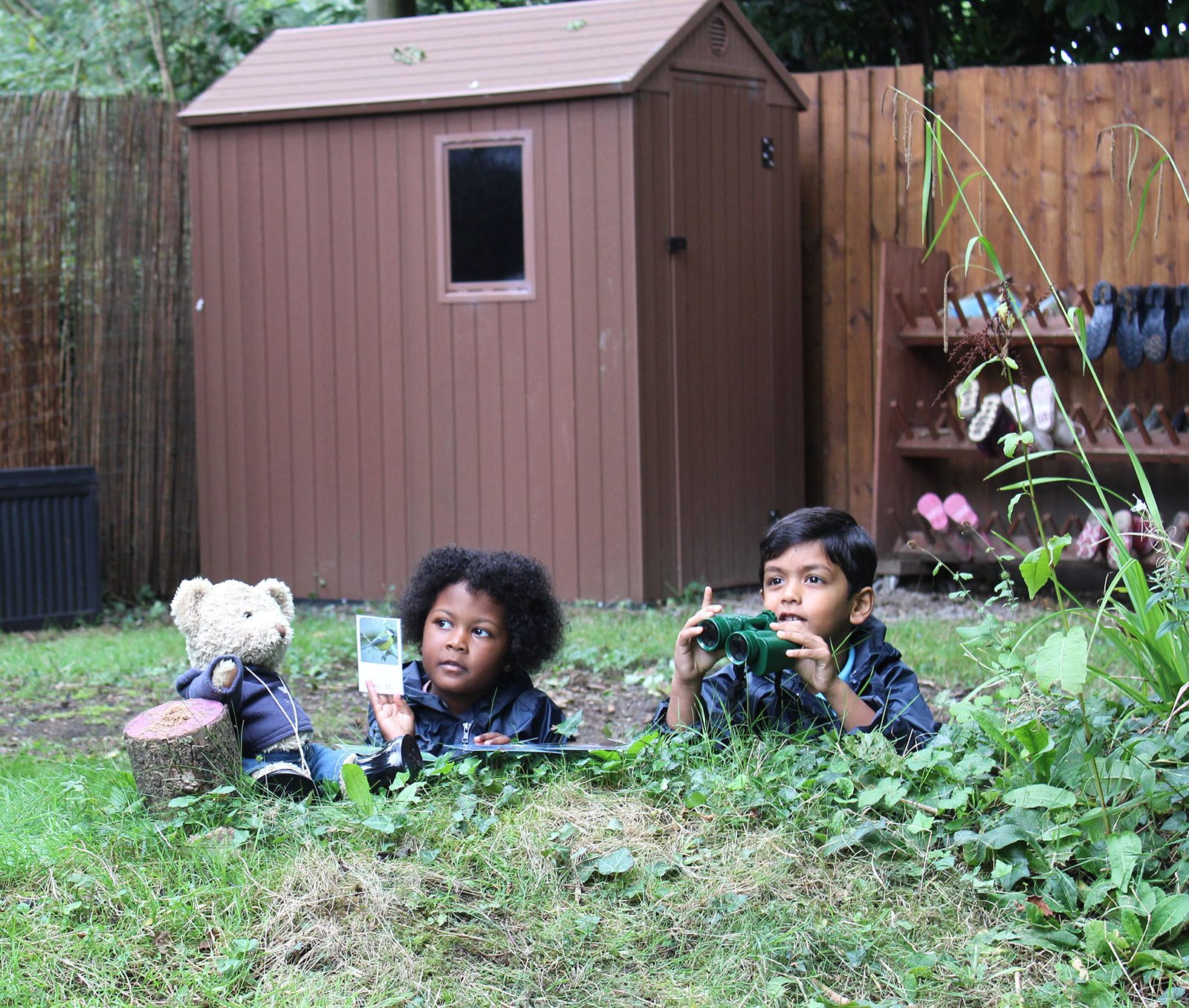 Acorns Nurseries Two children lie in the grass of Acorns Duffryn's backyard, one holding a book and the other with binoculars. A teddy bear sits beside them.