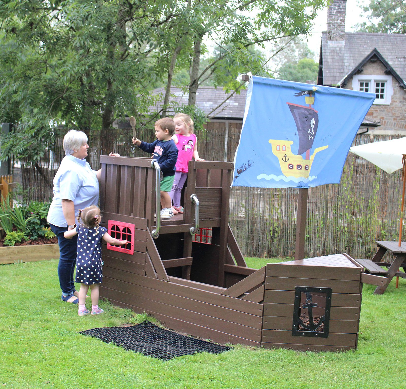 Acorns Nurseries Children are playing on a wooden pirate ship structure in the grassy area of Duffryn, supervised by an adult who occasionally gathers acorns for a spontaneous treasure hunt.