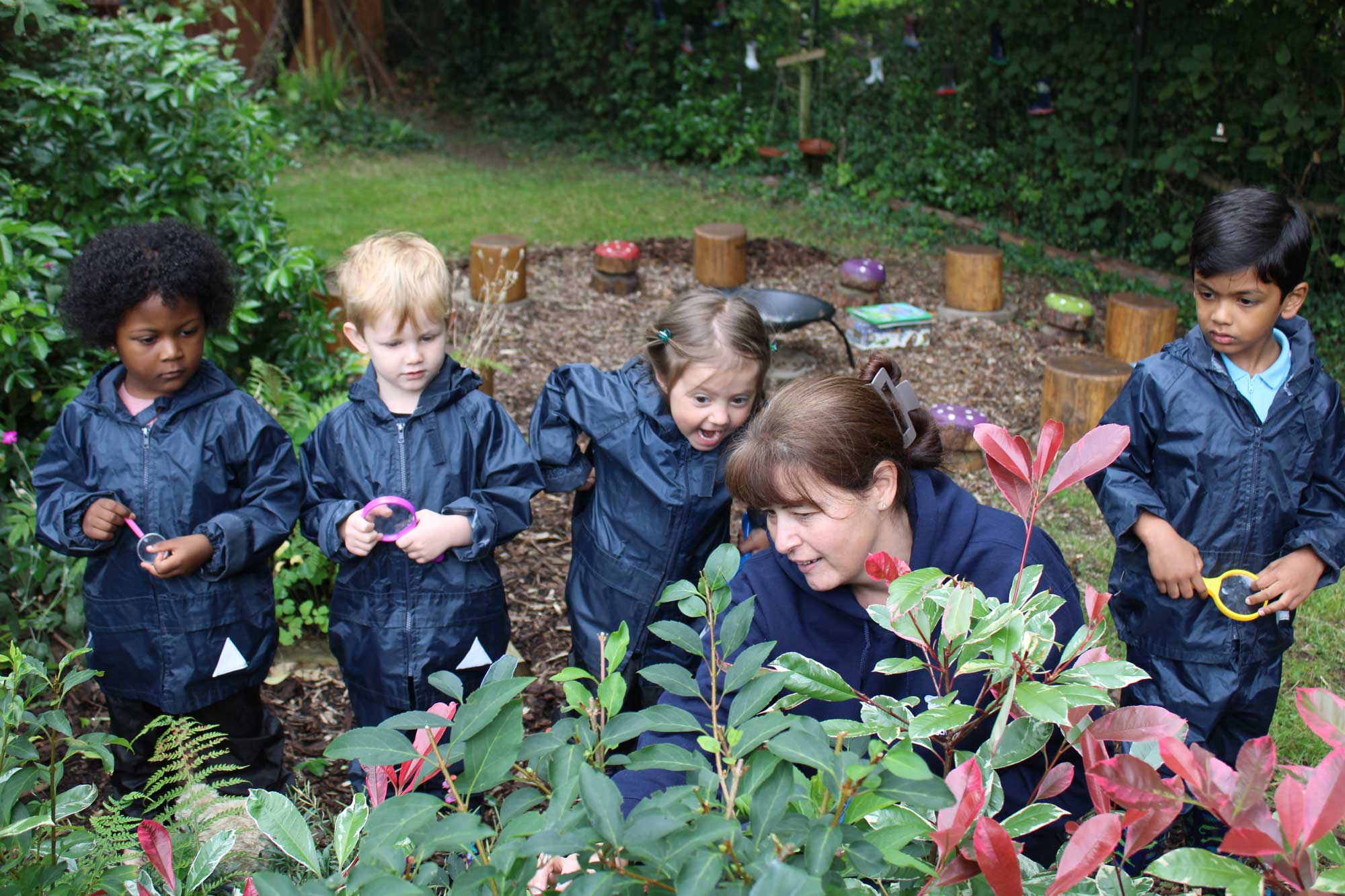 Acorns Nurseries Children in navy raincoats engage in outdoor education as they observe a woman teaching in a garden, surrounded by lush greenery and tree stumps.