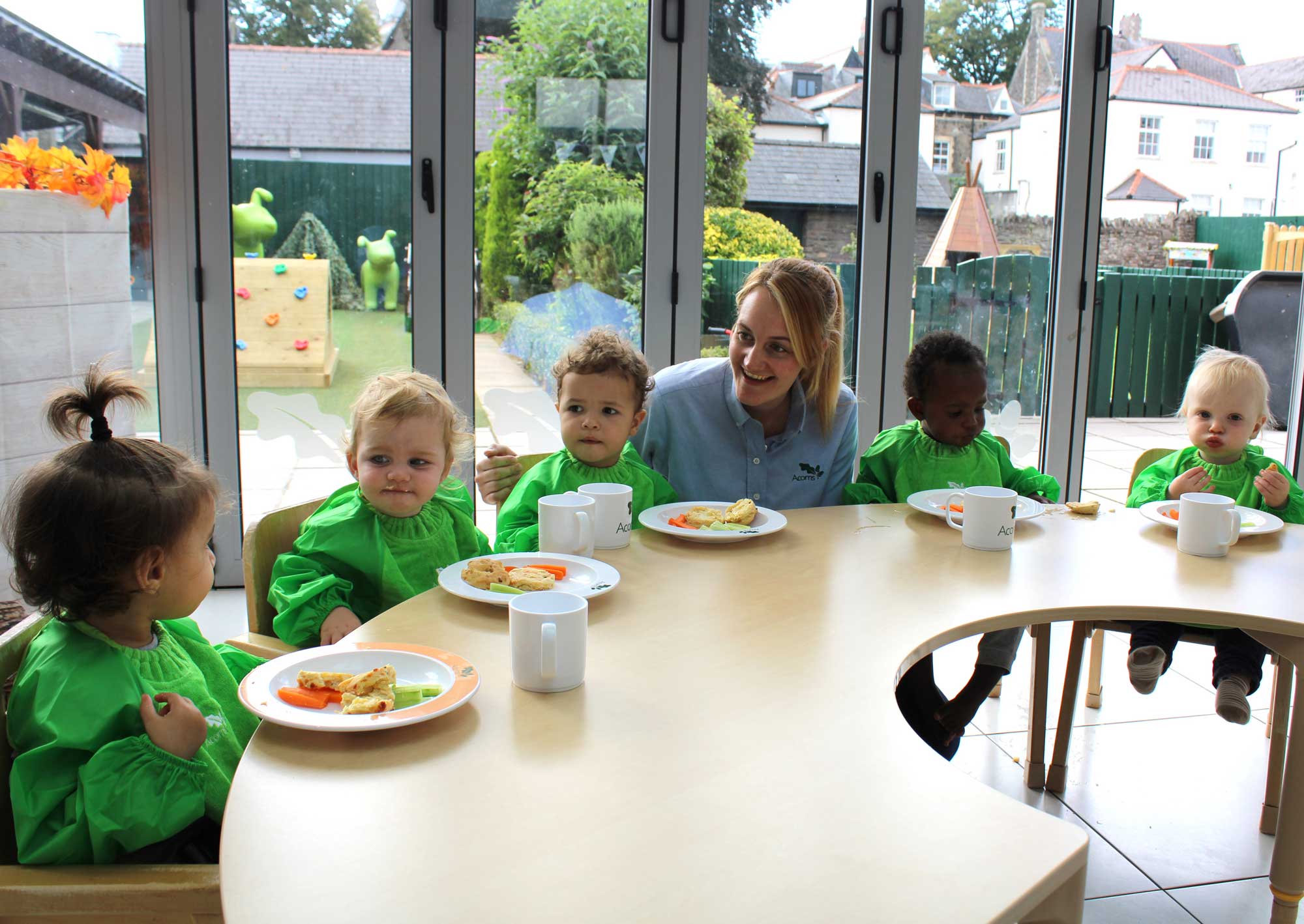 Acorns Nurseries Children in green smocks sit around a table with plates of nutritious food, accompanied by an adult, in a bright room with large windows.