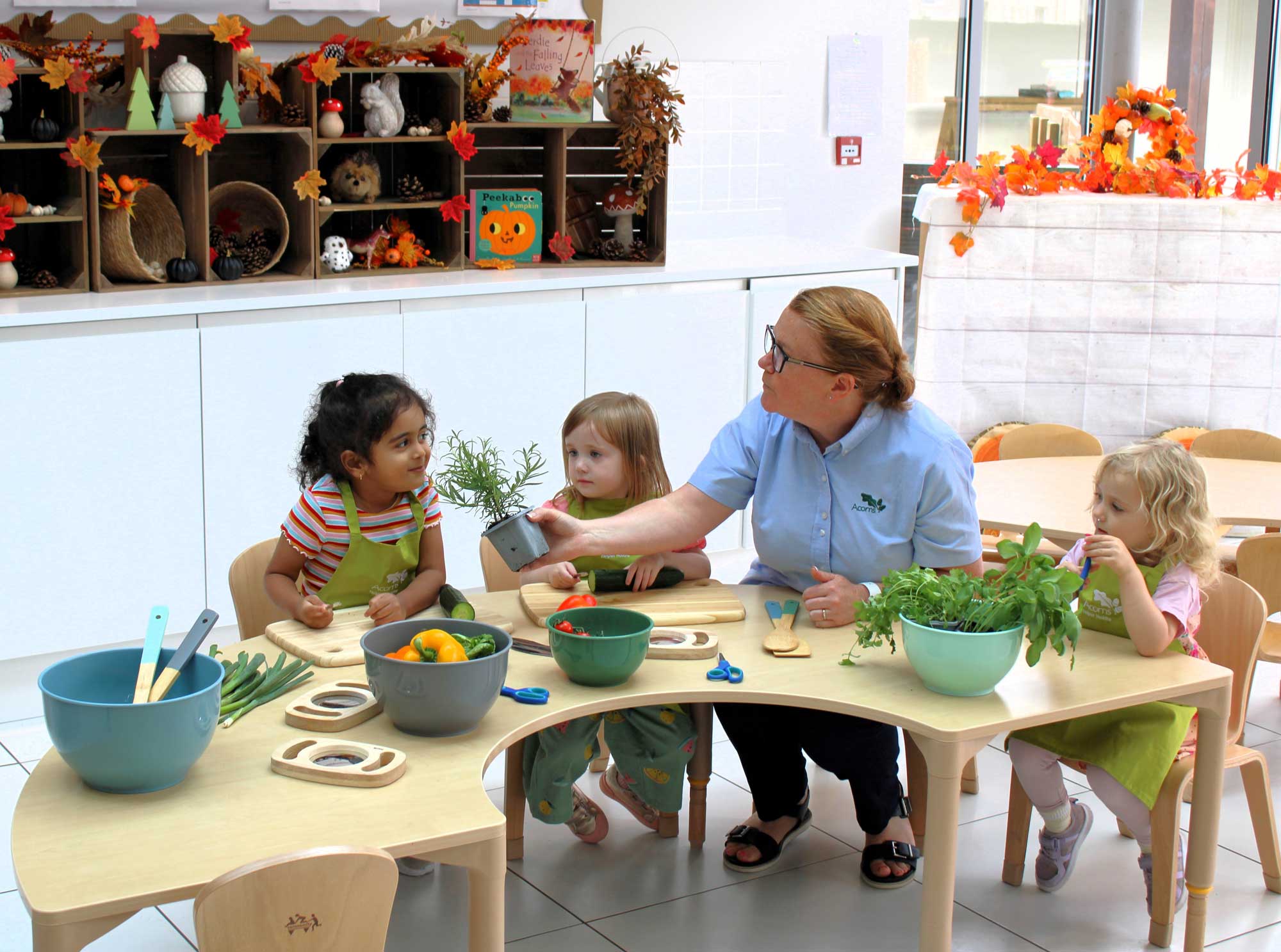 Acorns Nurseries Four children and a teacher interact at a table with vegetables and craft materials in the vibrant environment of a nursery classroom.