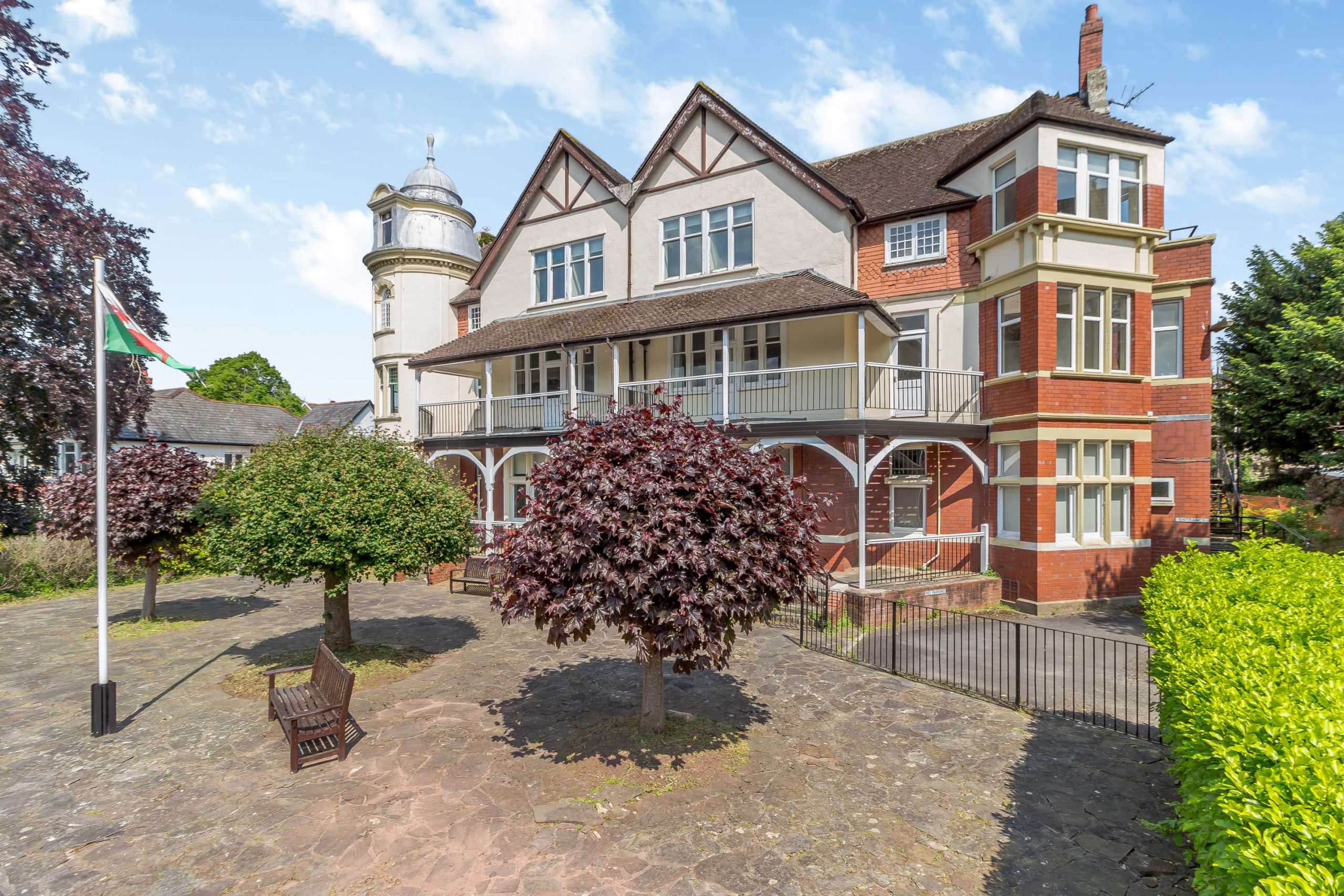 Acorns Nurseries Three-story building with red brick and white facade, surrounded by trees and courtyard with benches, flagpole on left.