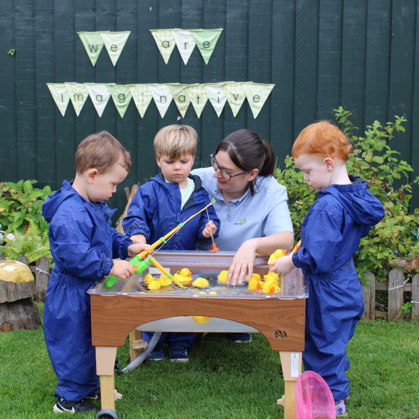 Acorns Nurseries A caregiver and three children, dressed in blue, play with rubber ducks at a water table outside, where their laughter echoes the joy of small investments growing like acorns turning into mighty oaks.