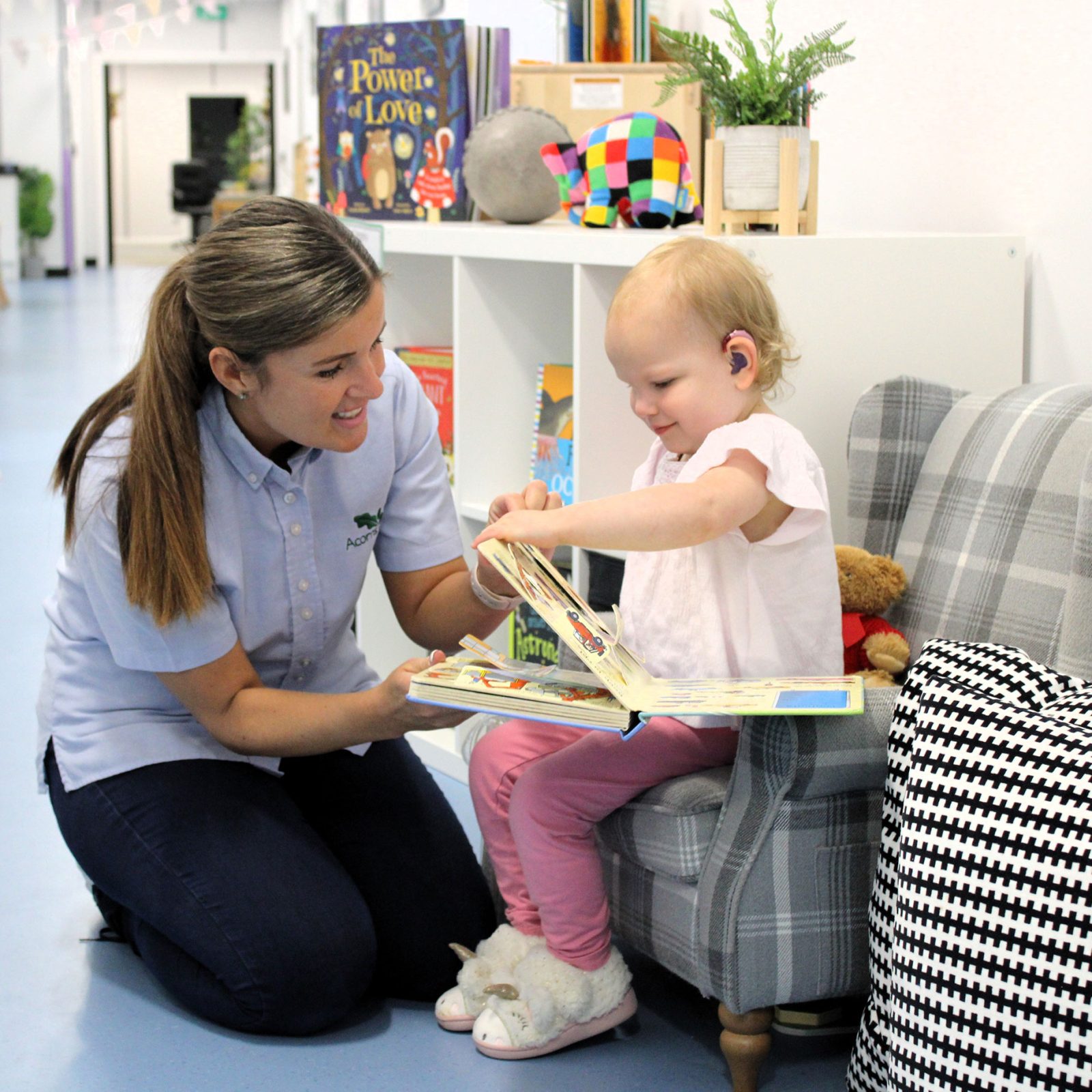 Acorns Nurseries A woman and a young child with hearing aids read a book together in a cozy room, surrounded by shelves of toys and books. Just like nurturing young minds, investing time in shared stories can plant acorns of knowledge that grow into lifelong treasures.