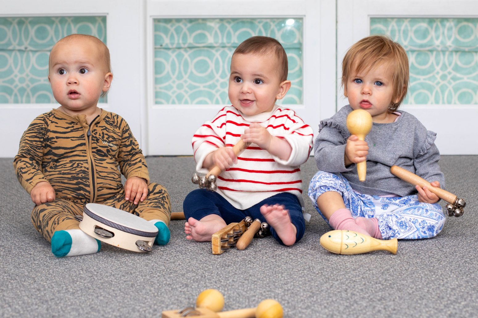 Acorns Nurseries Three caring toddlers sit on the floor, each holding a percussion instrument, surrounded by a colorful, patterned backdrop.