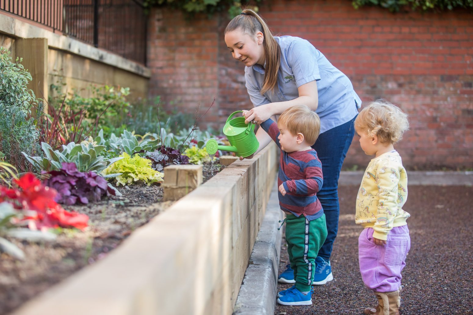 Acorns Nurseries A woman engages in children's care as she helps two young children water plants in a garden, nurturing their sense of responsibility and connection to nature. The children watch eagerly as she pours from a green watering can, fostering their well-being and curiosity.