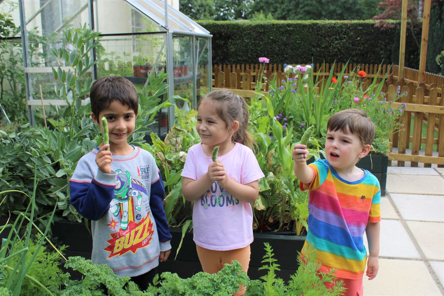 Acorns Nurseries Three children holding fresh vegetables stand in a lush garden, smiling in front of plants and a greenhouse, celebrating the joy of healthy eating.