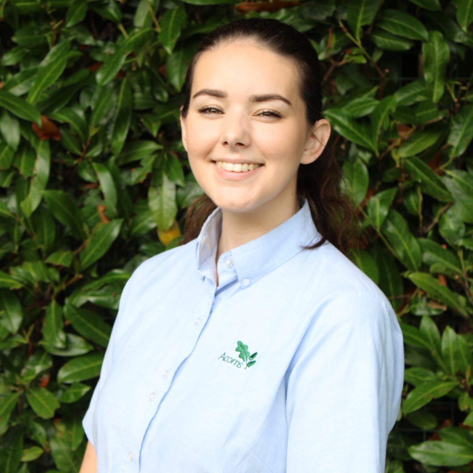 Acorns Nurseries Person in a light blue shirt with a green logo, standing in front of a leafy background, smiling at the camera, exemplifying collaboration across teams.