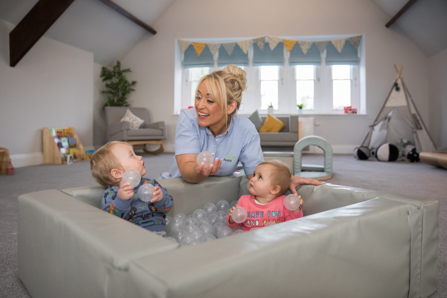 Acorns Nurseries A caregiver smiles at two babies playing with plastic balls in the nursery's padded play area, creating a nurturing environment inside a spacious room with large windows.