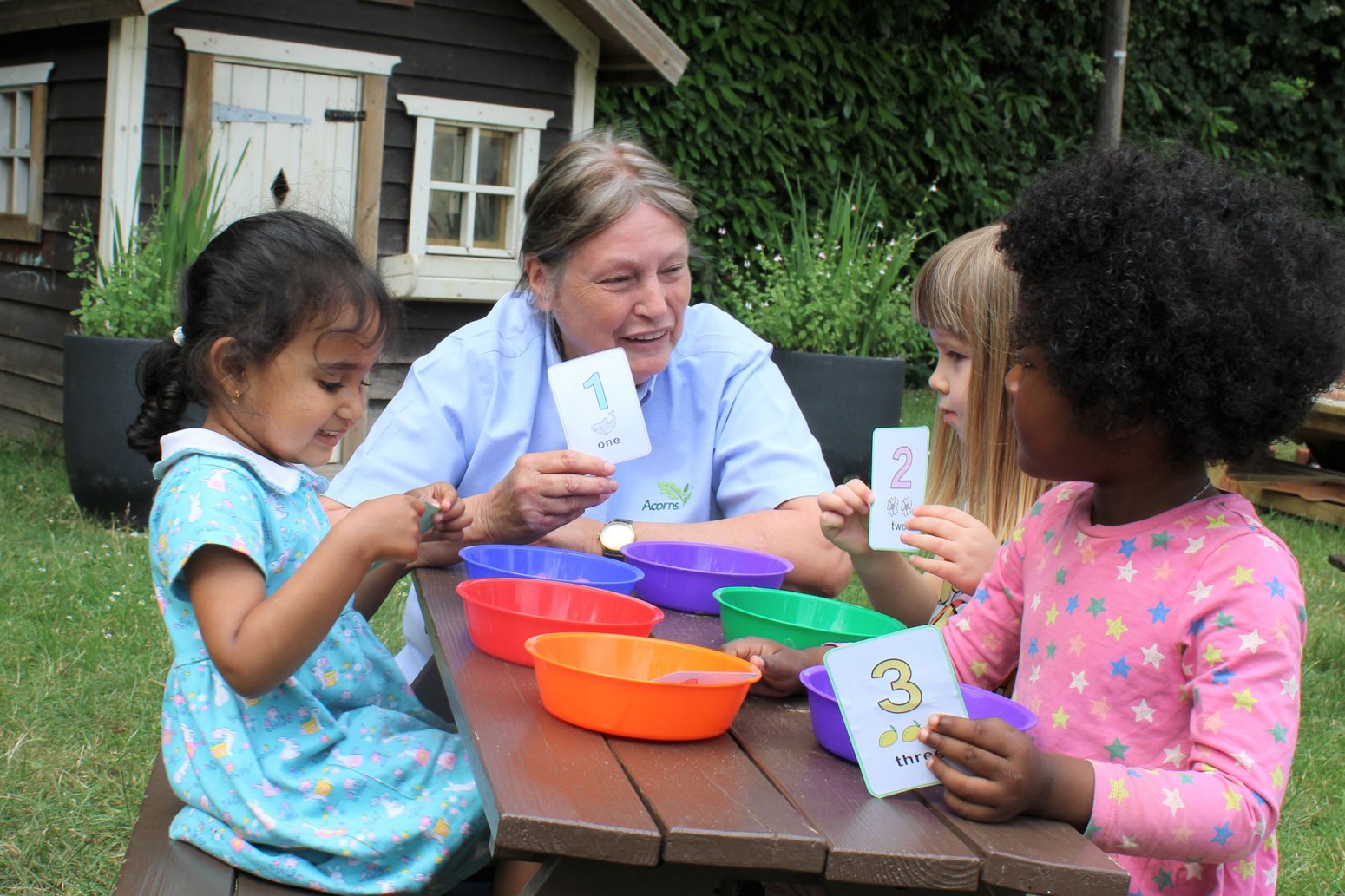 Acorns Nurseries At a picnic table nestled in the Acorns garden, a teacher engages three curious children in learning numbers with cards and bowls, creating an enriching experience for those who've already completed child registration.