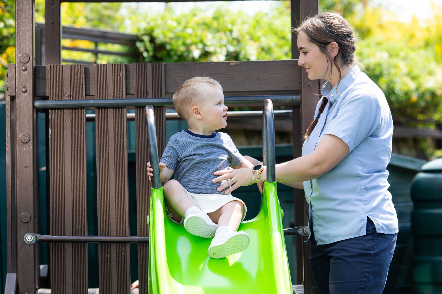 Acorns Nurseries A woman helps a child seated at the top of a green slide at a playground, surrounded by wooden play structures and scattered acorns in the greenery.