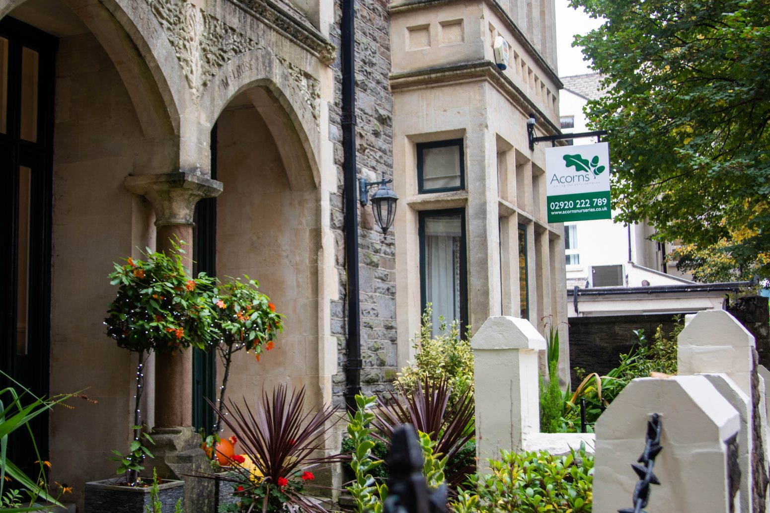 Acorns House - Acorns Nurseries A stone building with arched windows stands proudly, displaying a sign for Acorns above its charming, gated garden filled with shrubs and a small tree. This picturesque location invites visitors to enjoy its serene ambiance.