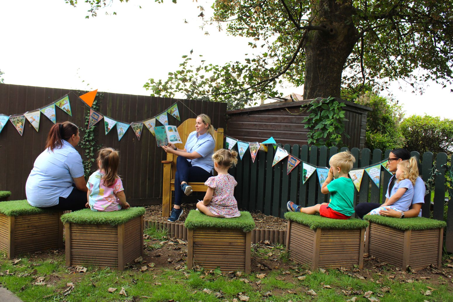 Acorns Nurseries Outdoor storytime session with two adults and four children, surrounded by acorns, seated on wooden benches near trees and a decorated fence.