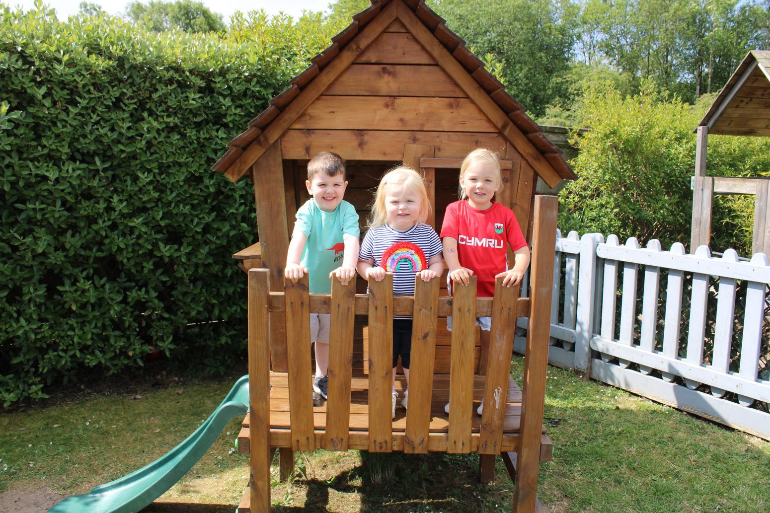 Acorns Nurseries Three children stand smiling on the porch of a wooden playhouse, surrounded by lush greenery and scattered acorns, against a backdrop of a garden fence.