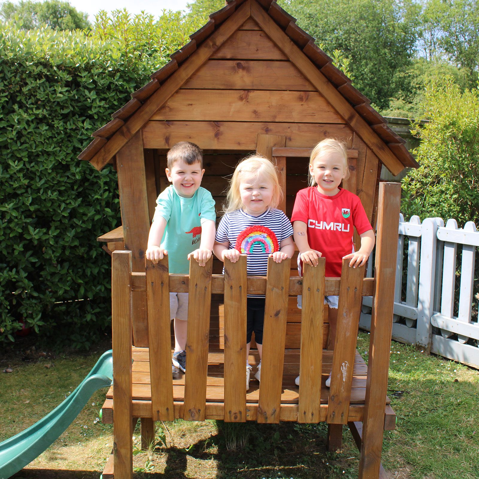 Acorns Nurseries Three children stand smiling on the porch of a wooden playhouse, surrounded by lush greenery and scattered acorns, against a backdrop of a garden fence.