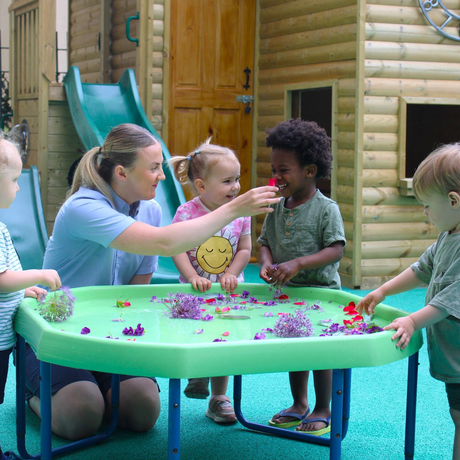 Acorns Nurseries Children and a caregiver enjoy a playful afternoon, exploring acorns and flowers together at the water table in an outdoor play area with a slide and playhouse.