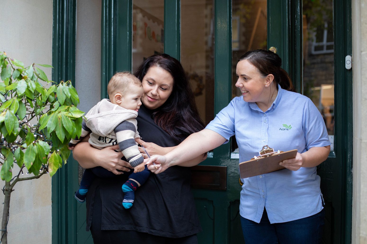 Acorns Nurseries A woman holding a baby interacts with another woman registering details on a clipboard in front of a doorway adorned with a small potted acorn plant.