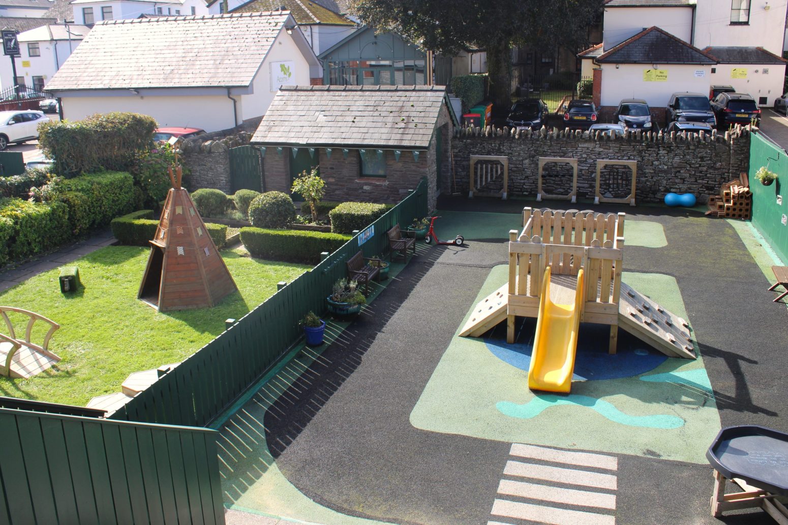 Acorns Nurseries An aerial view of a nursery playground showcases slides, a playhouse, and grassy areas, all embraced by a fence and nestled among nearby residential buildings, creating a nurturing environment.