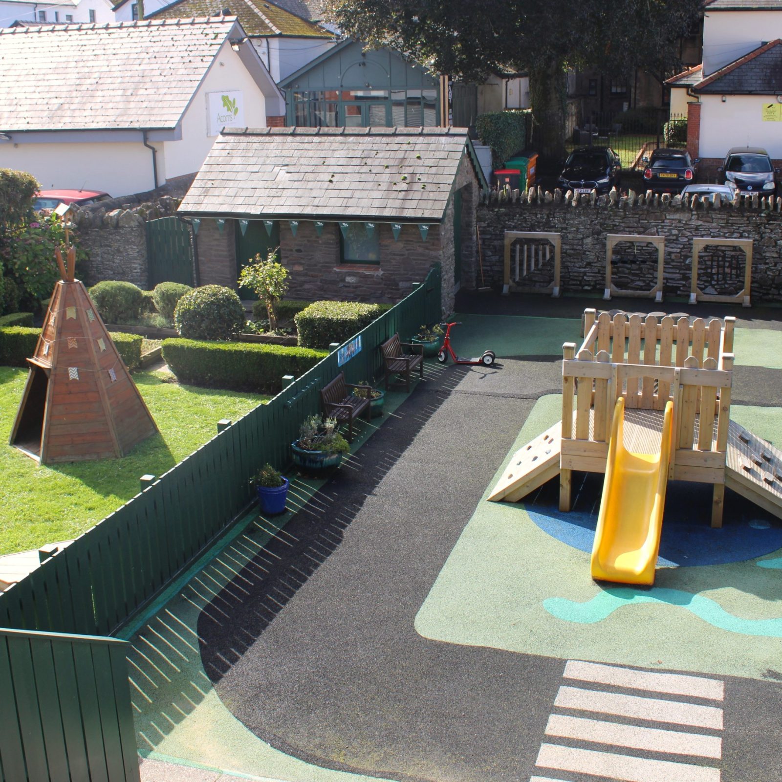 Acorns Nurseries A small playground with a slide, play structures, and a grass area offers children the joy of the Great Outdoors. Enclosed by a green fence and stone wall, this space includes a wooden shed for extra adventures.