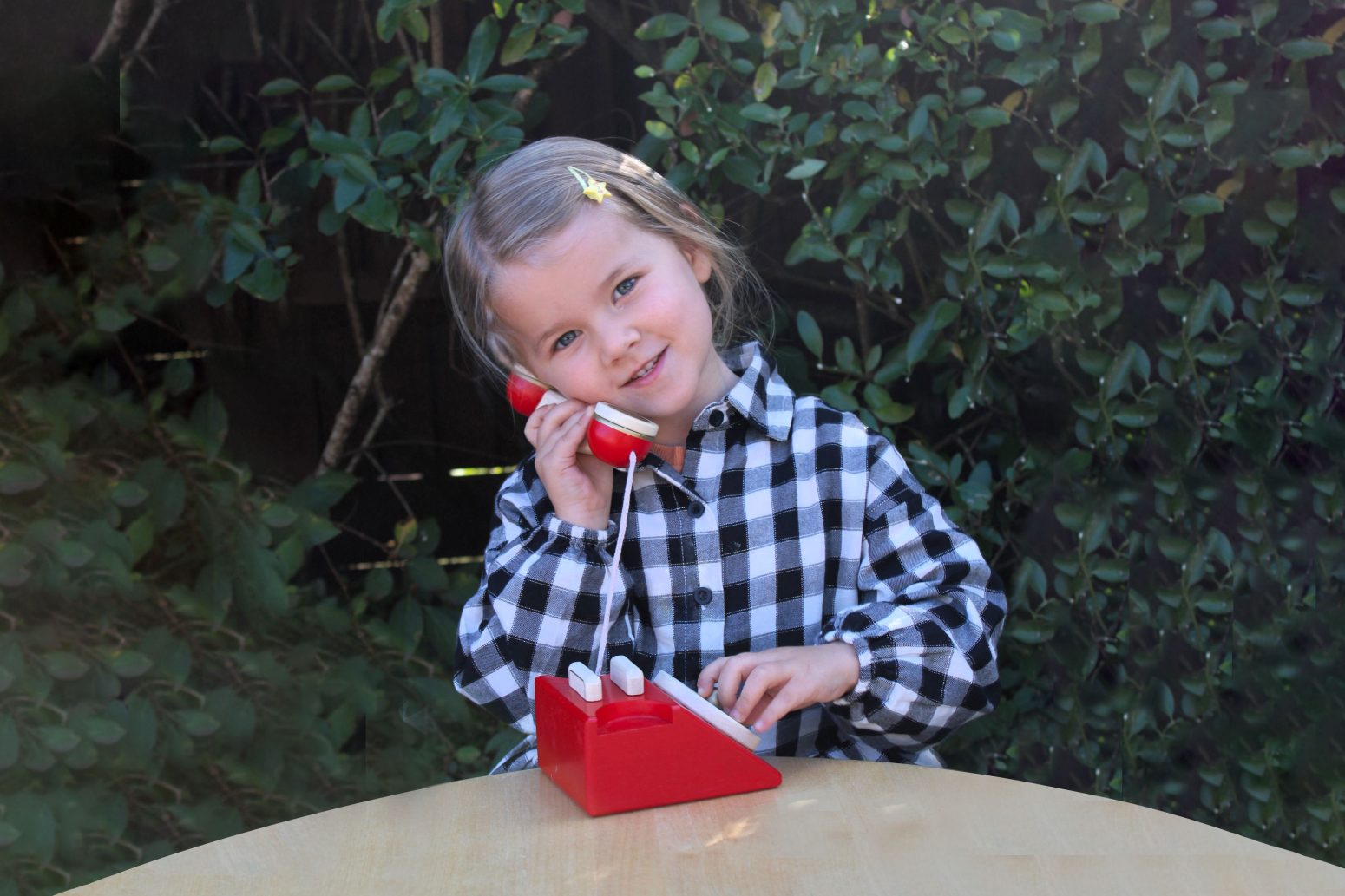 Acorns Nurseries A child in a checkered shirt playfully pretends to contact us using a toy phone, seated at a wooden table with lush foliage in the background.