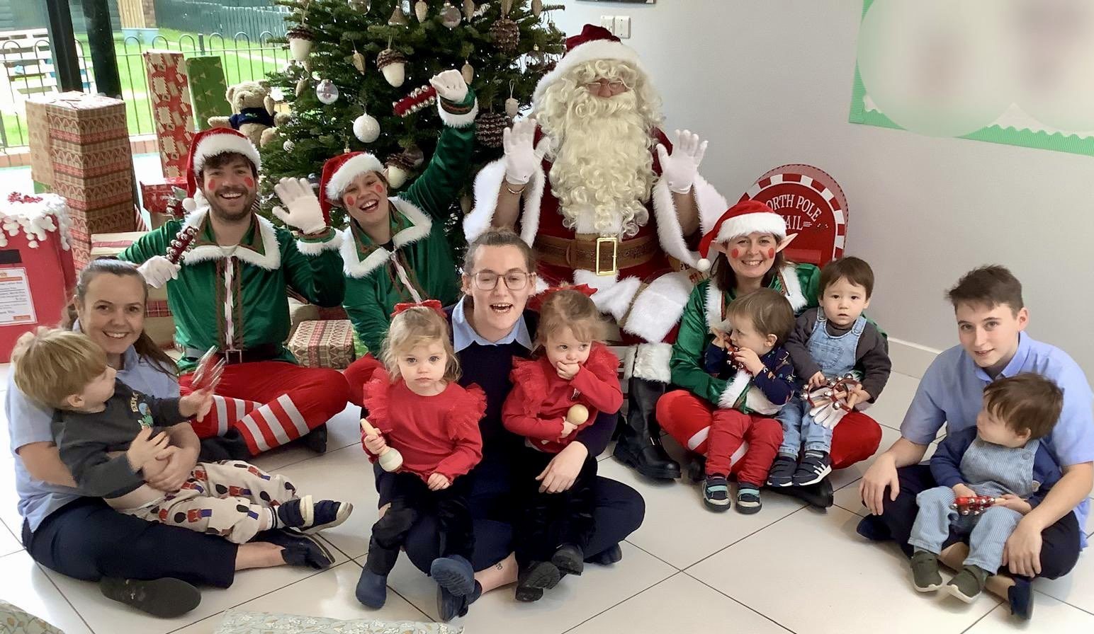 Acorns Nurseries Group of people with children from nurseries sitting on the floor in front of a decorated Christmas tree with Santa and elves.