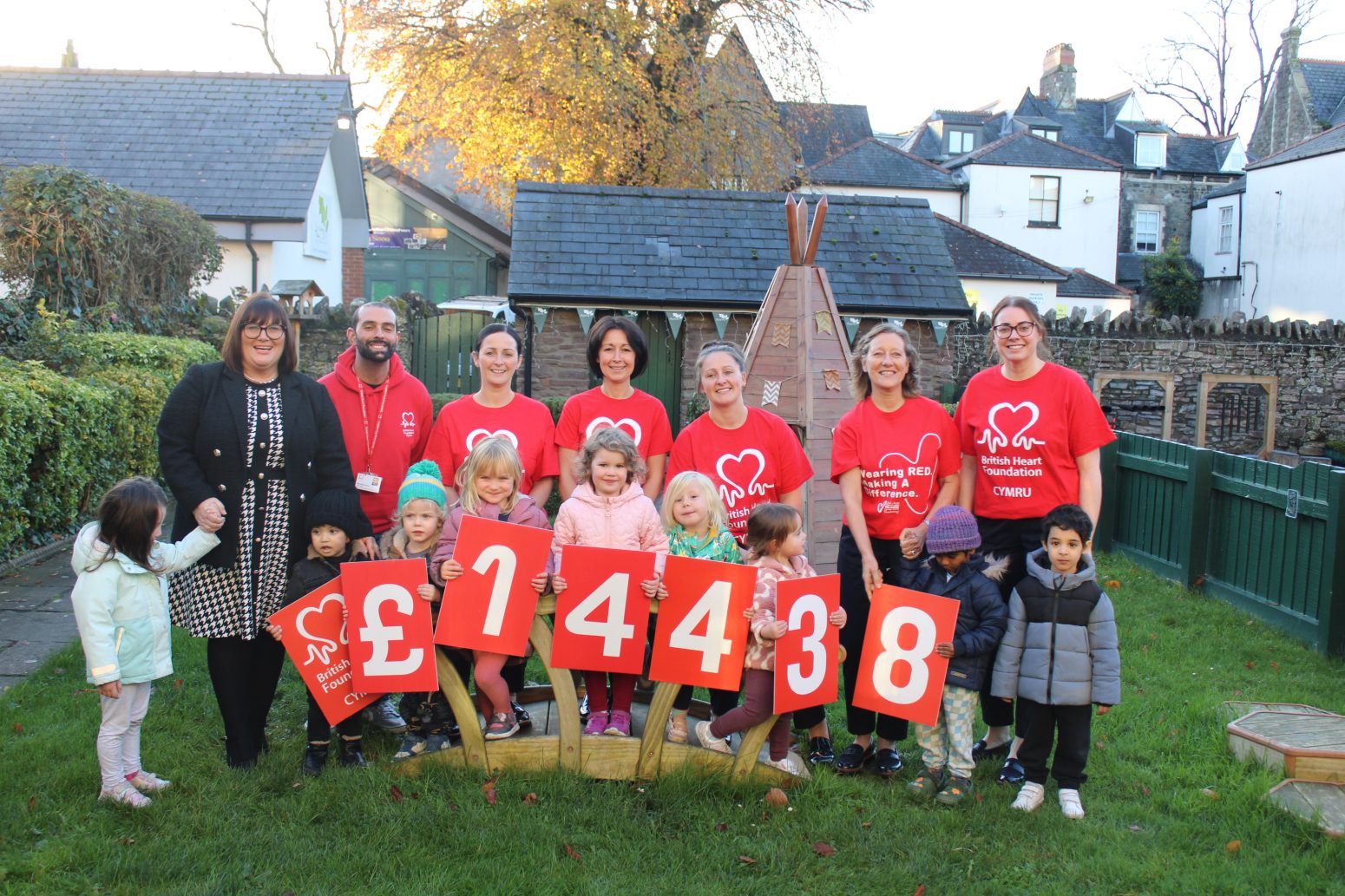 Acorns Nurseries A group of adults and children display large cards showing £14,438, standing outside in a garden area.