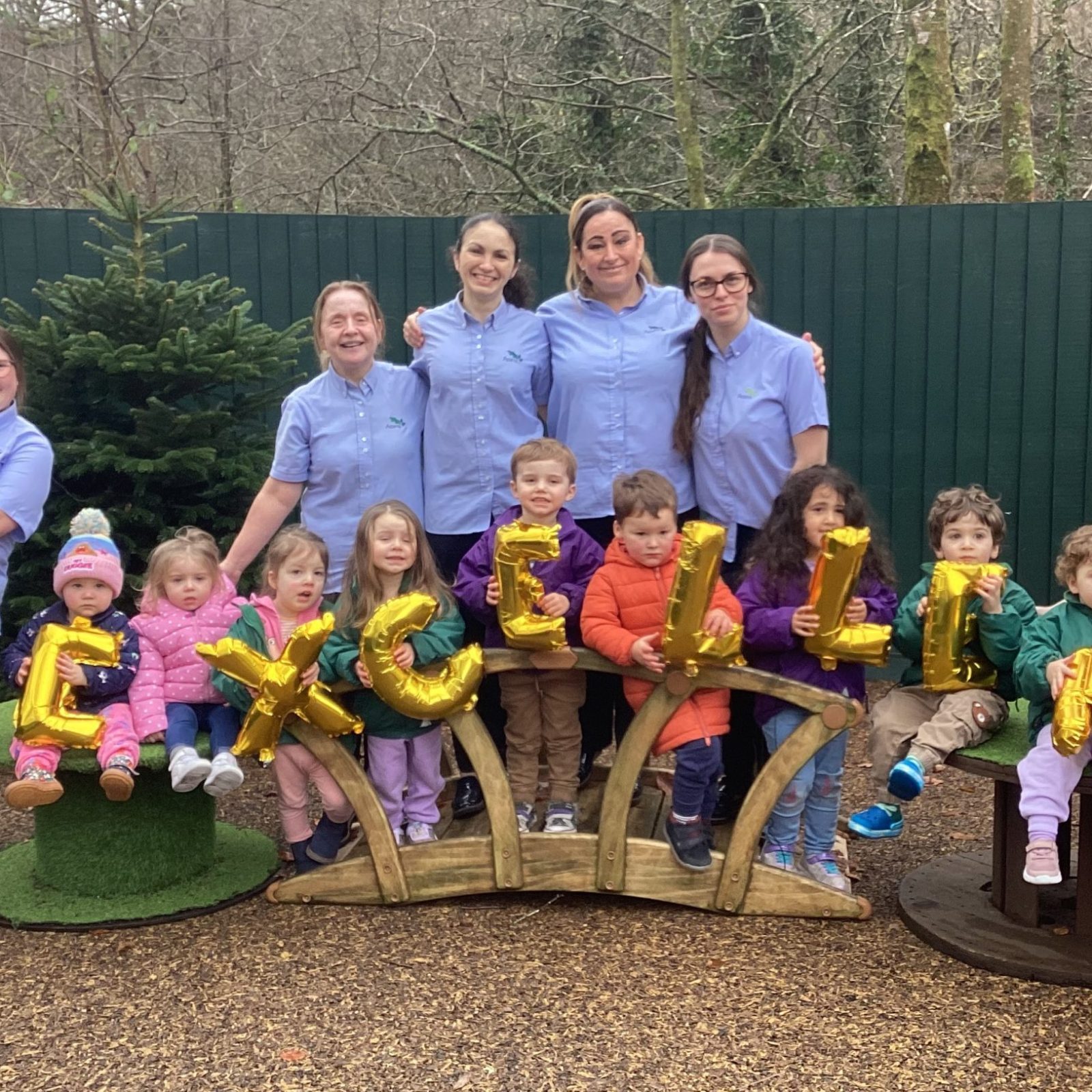 Acorns Nurseries A group of adults and children, including USW members, pose outdoors holding gold balloons spelling "EXCELLENT" in front of a fence and trees, celebrating a successful inspection.