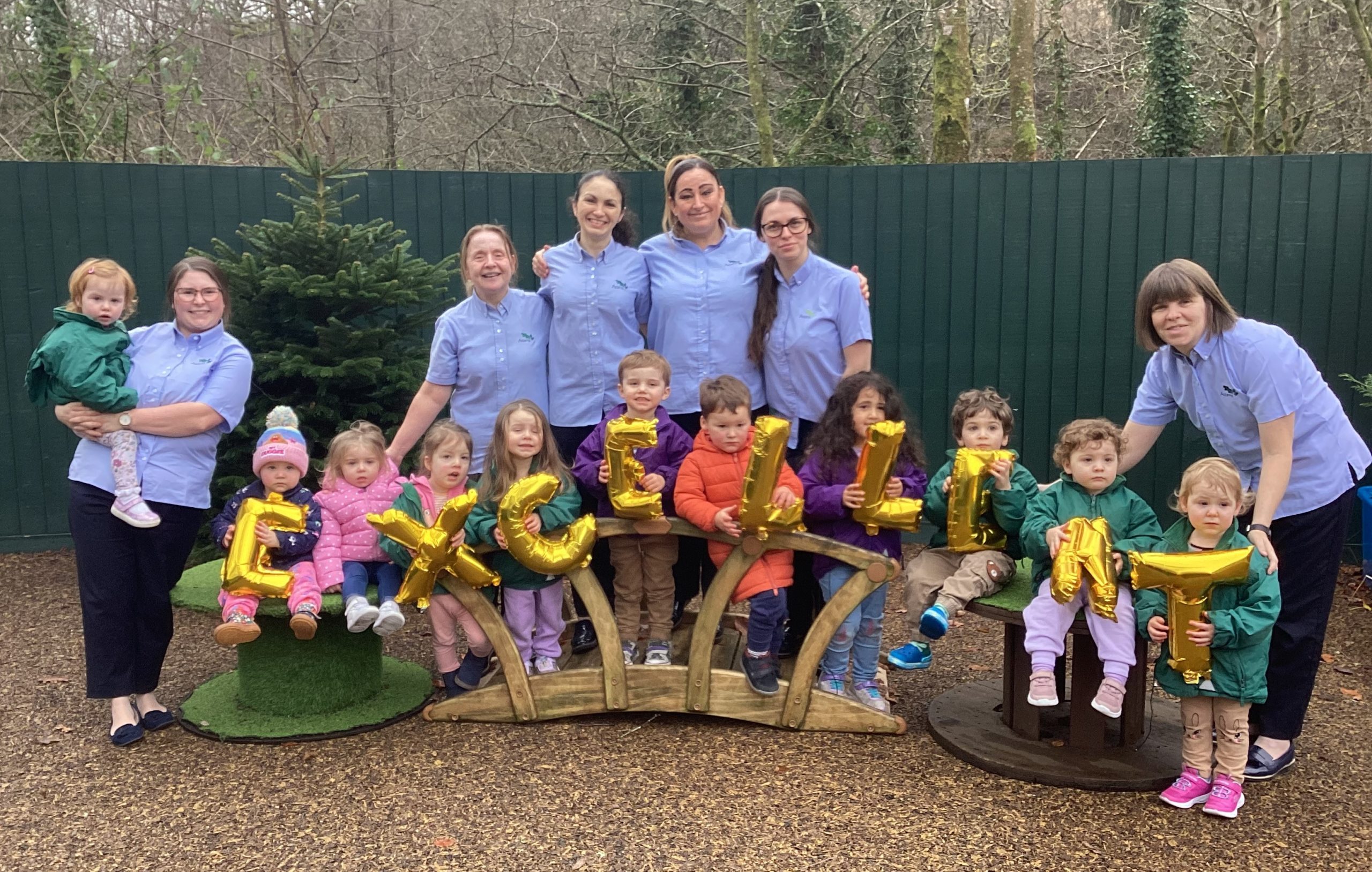 Acorns Nurseries A group of adults and children, including USW members, pose outdoors holding gold balloons spelling "EXCELLENT" in front of a fence and trees, celebrating a successful inspection.