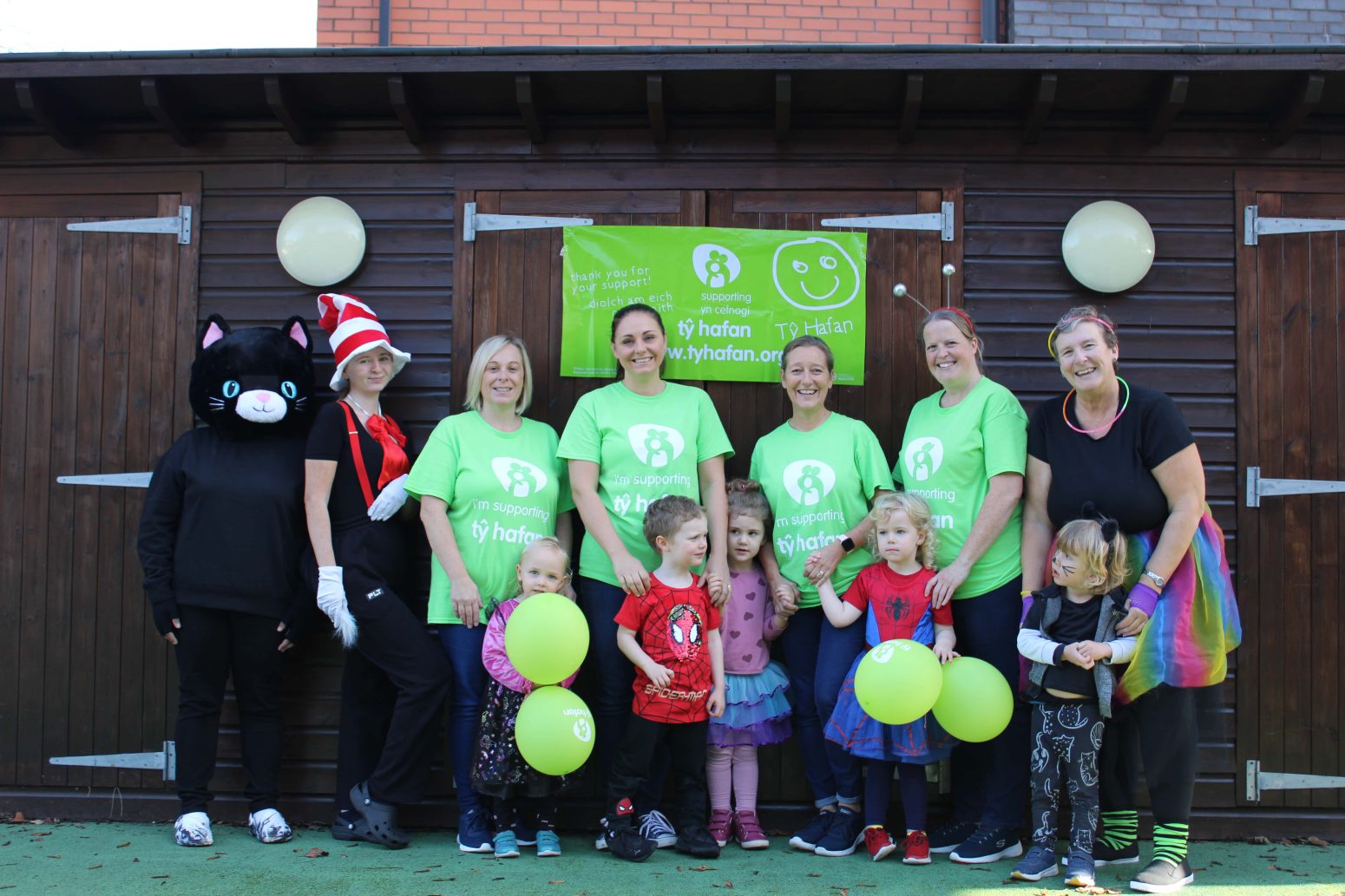 Acorns Nurseries Adults and children in costumes and green shirts pose with balloons in front of a wooden building, showcasing community involvement.